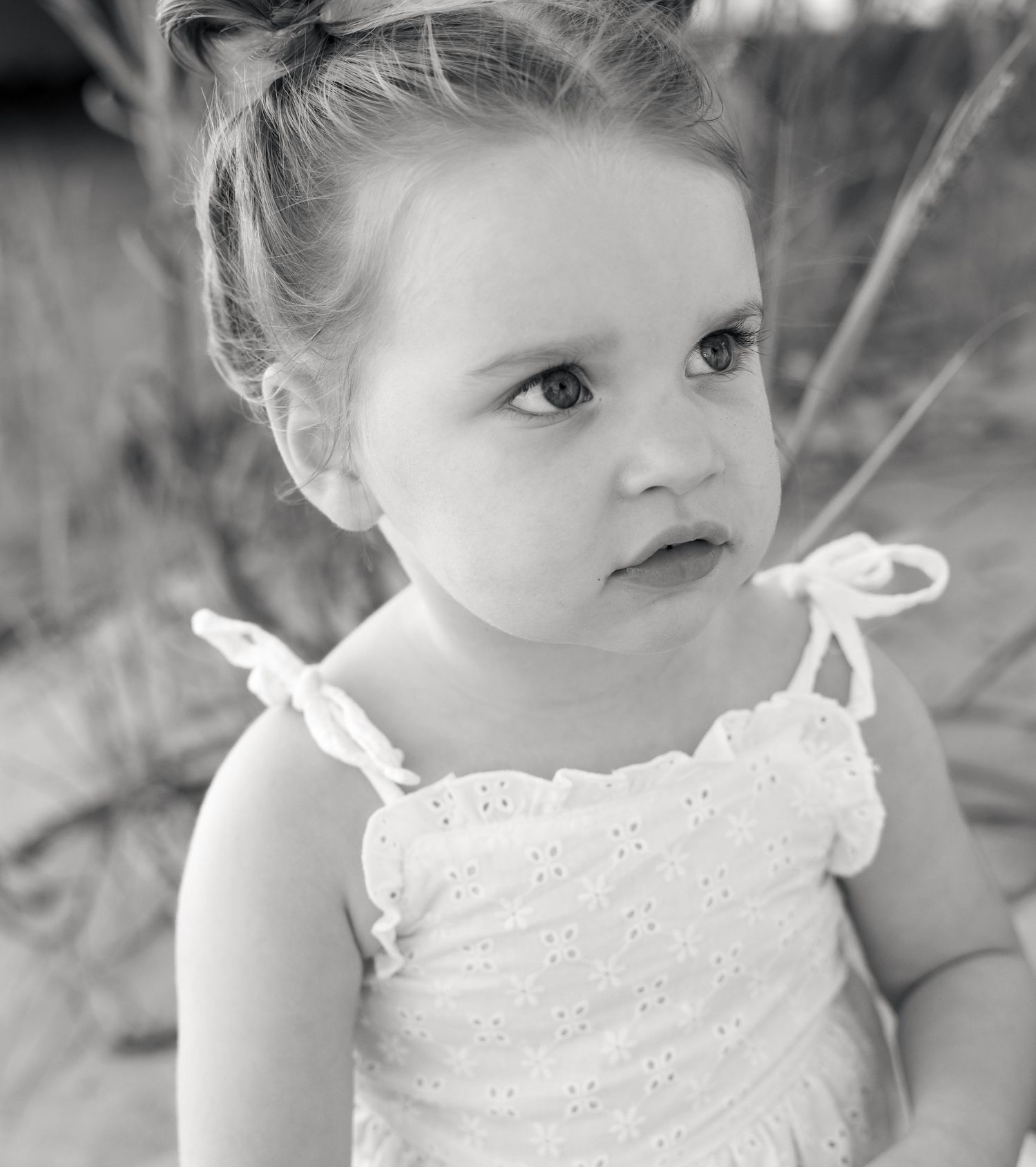 Black and white portrait of a toddler girl with hair in a top knot, wearing an eyelet sundress with tied shoulder straps outdoors.