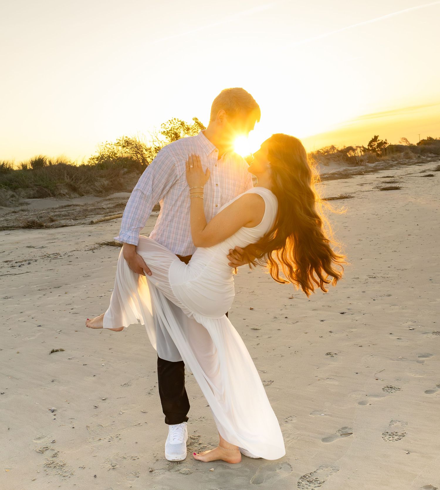 Couple sharing a romantic dip kiss on sandy beach at golden sunset, woman in white dress, man in plaid shirt.