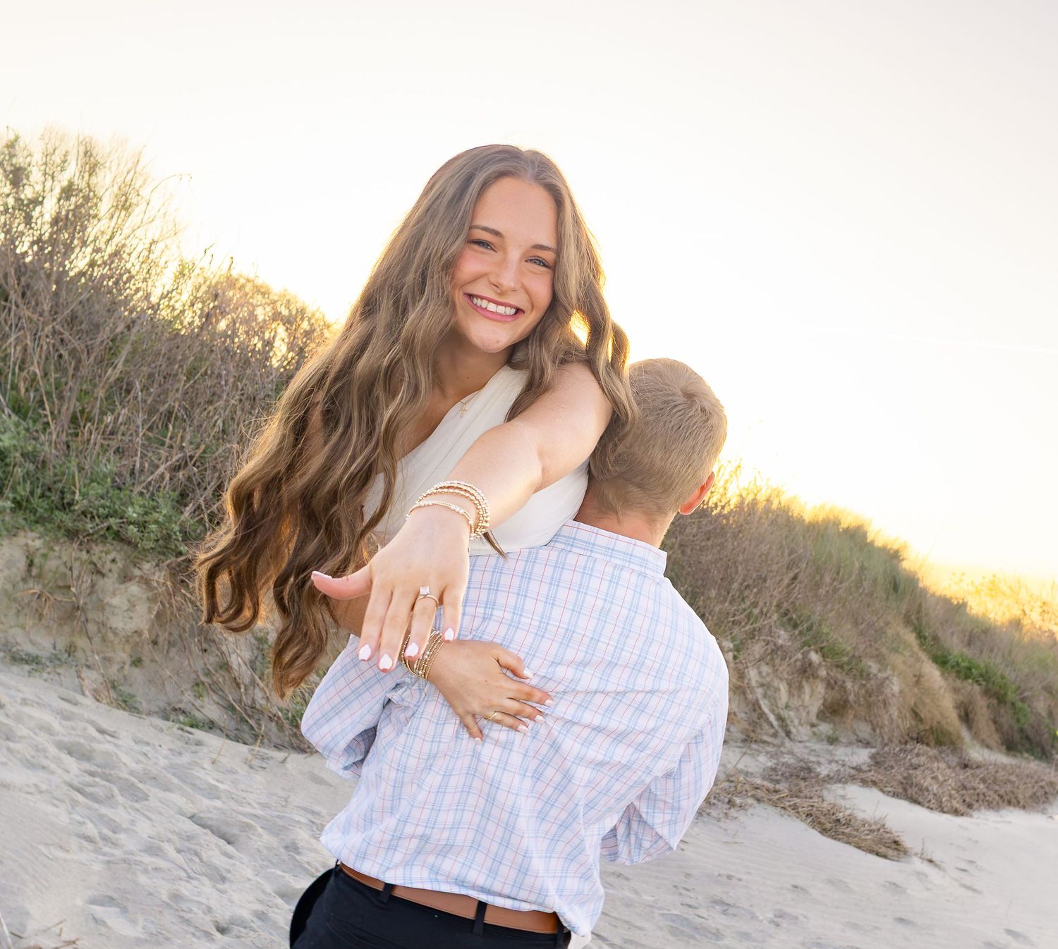 Couple embracing on a sandy beach at sunset, woman with long wavy hair smiling and showing engagement ring.