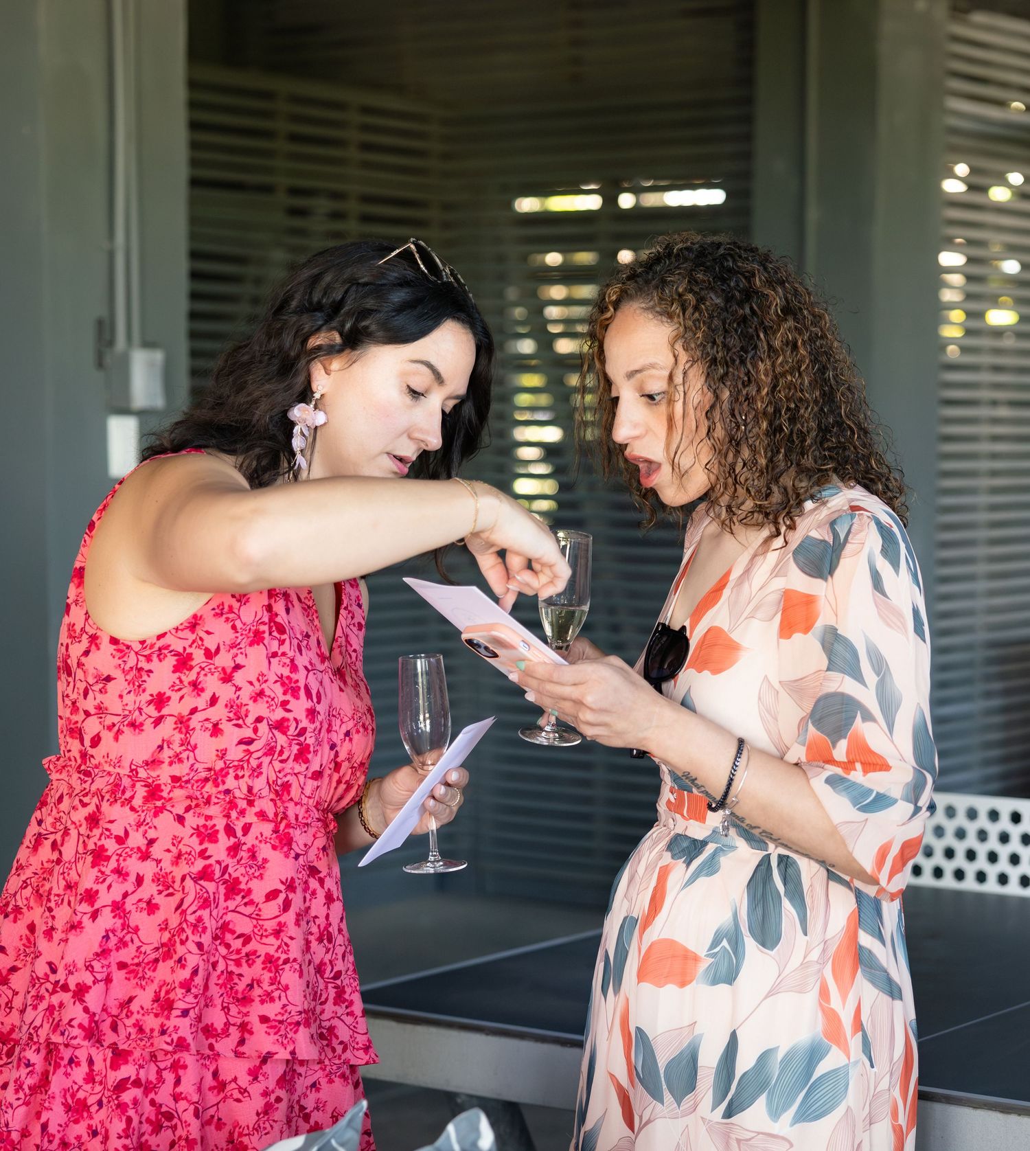 Two women in floral dresses reviewing a card while holding champagne glasses outdoors.