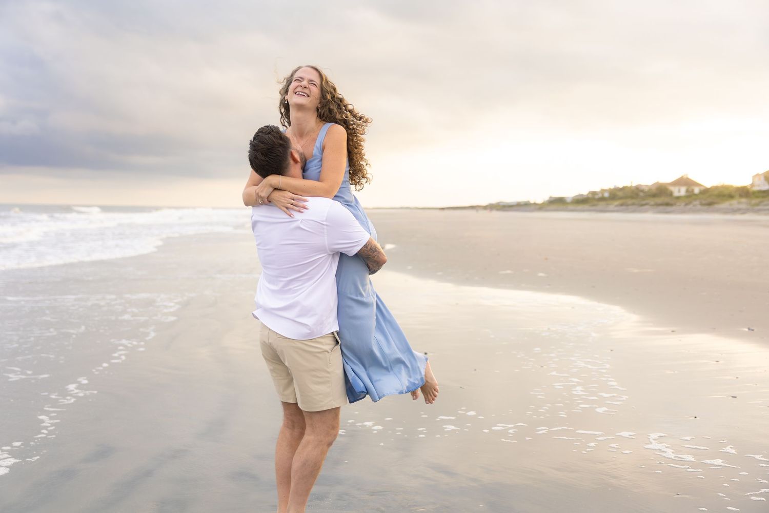 Couple embracing on a beach at sunset, woman laughing as man lifts her in a romantic seaside moment.