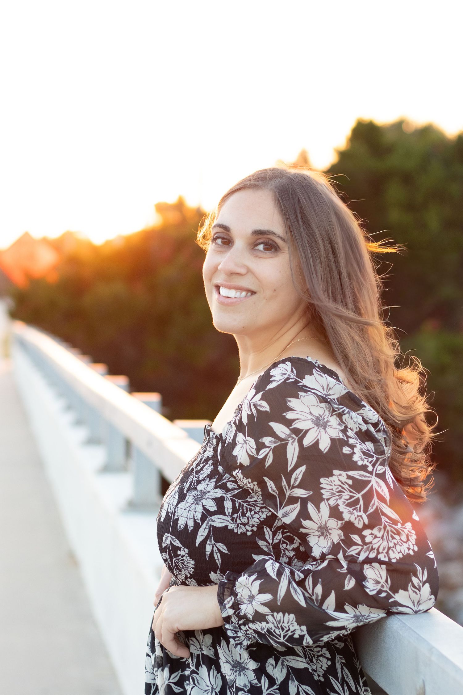 Woman in floral dress smiling on bridge at golden hour sunset with trees in background.