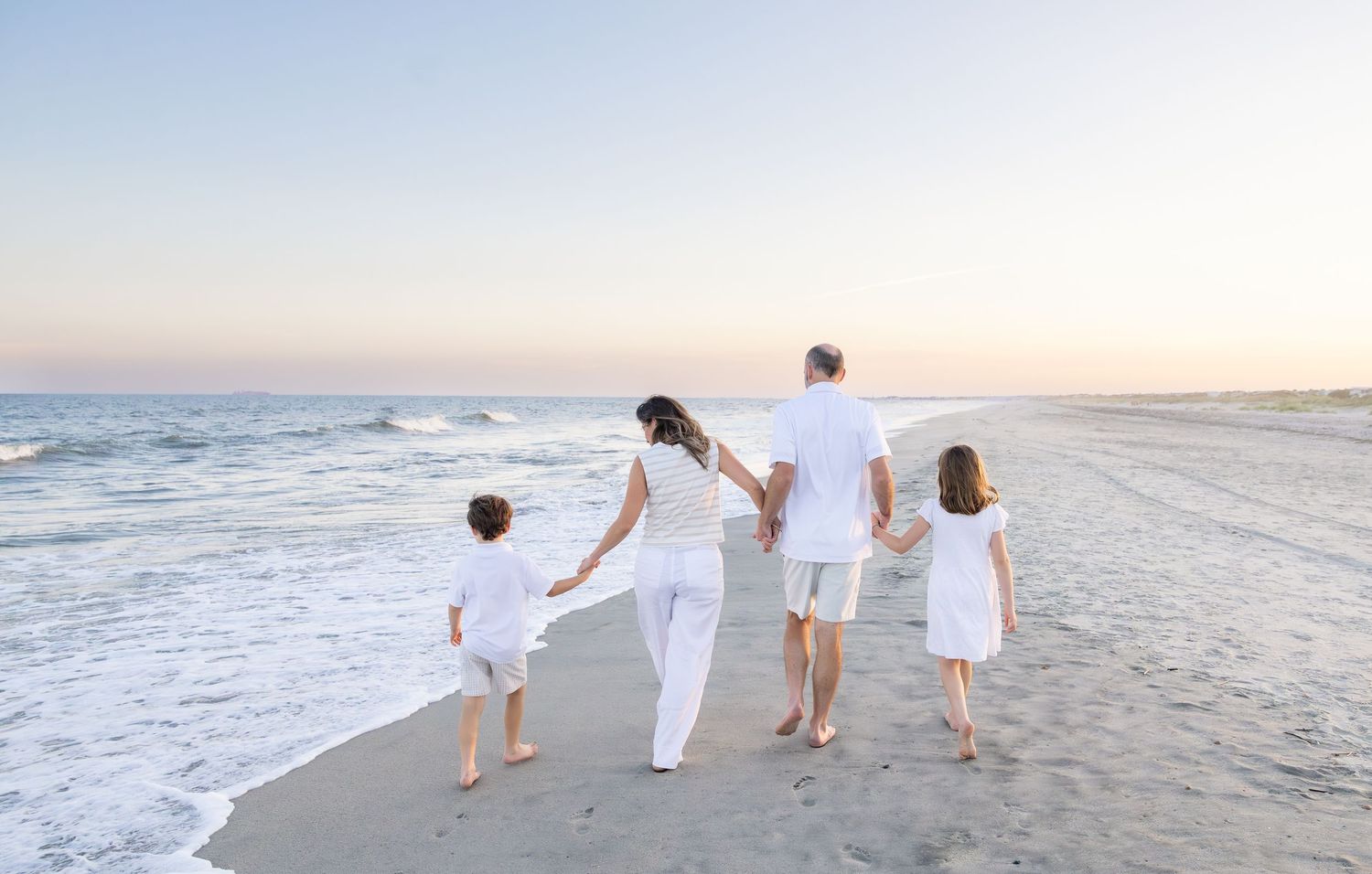 Family of four in white outfits walking hand-in-hand along a serene beach at sunset.