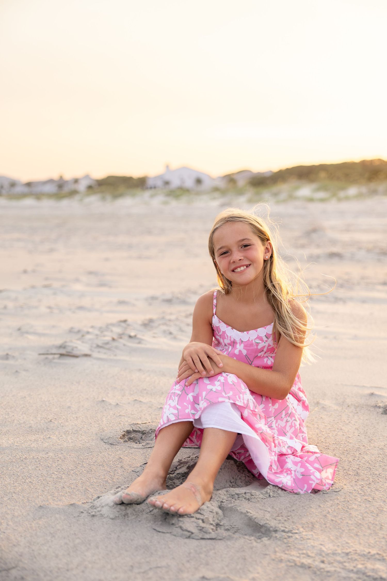 Young girl in pink floral dress sitting barefoot on sandy beach at sunset, smiling at camera.