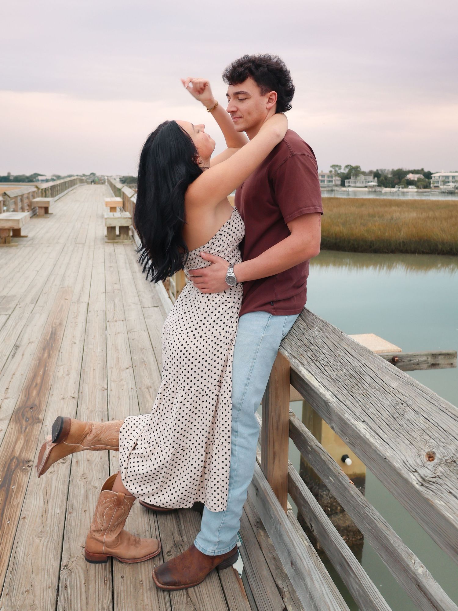 Couple dancing on a wooden pier at sunset, woman in polka dot dress and cowboy boots, man in burgundy shirt and jeans.