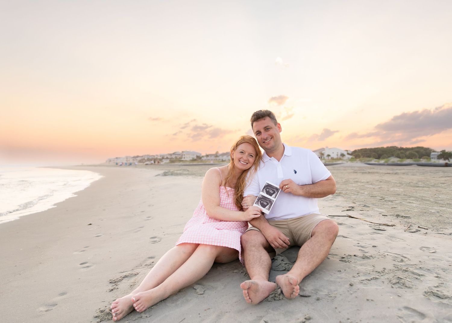 Couple sitting on sandy beach at sunset holding ultrasound photos, announcing pregnancy with pastel sky backdrop.