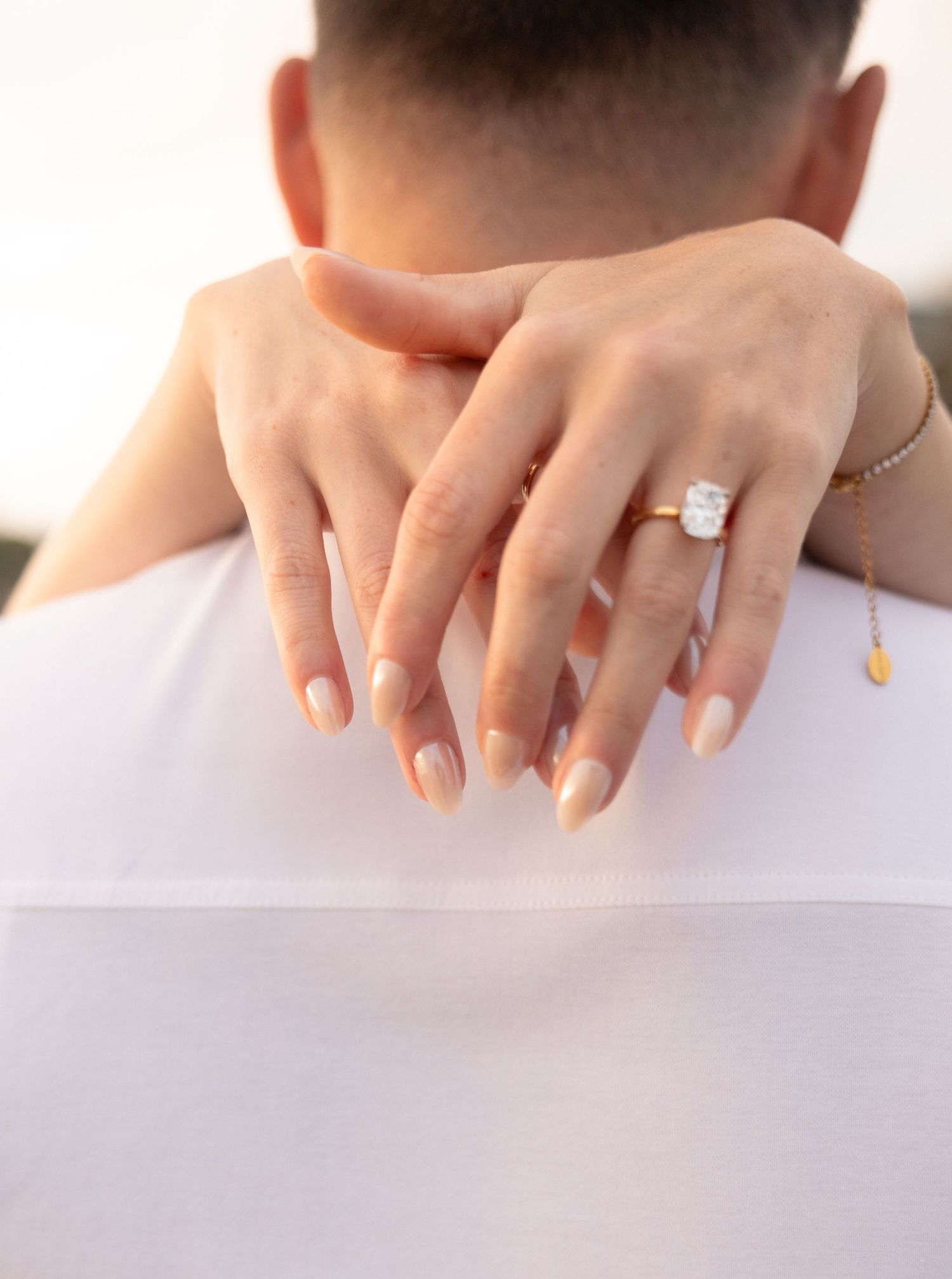 Close-up of a woman's hand with nude nails and a diamond engagement ring resting on a man's shoulder.