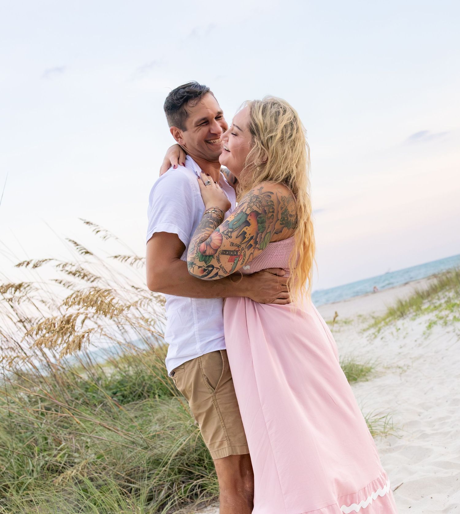 A couple embraces and smiles on a sandy beach, the woman wearing a pink dress with tattoos, sea oats in the background.