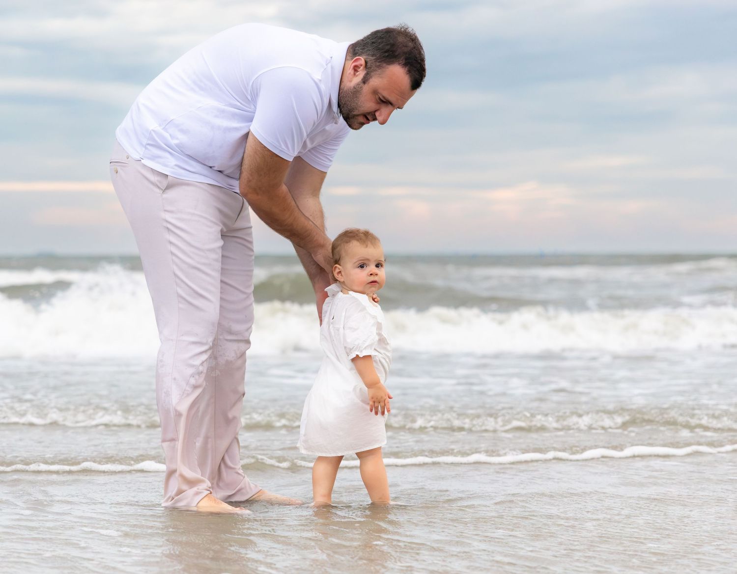 Father helping toddler walk on sandy beach near ocean waves, both dressed in white on a cloudy day.