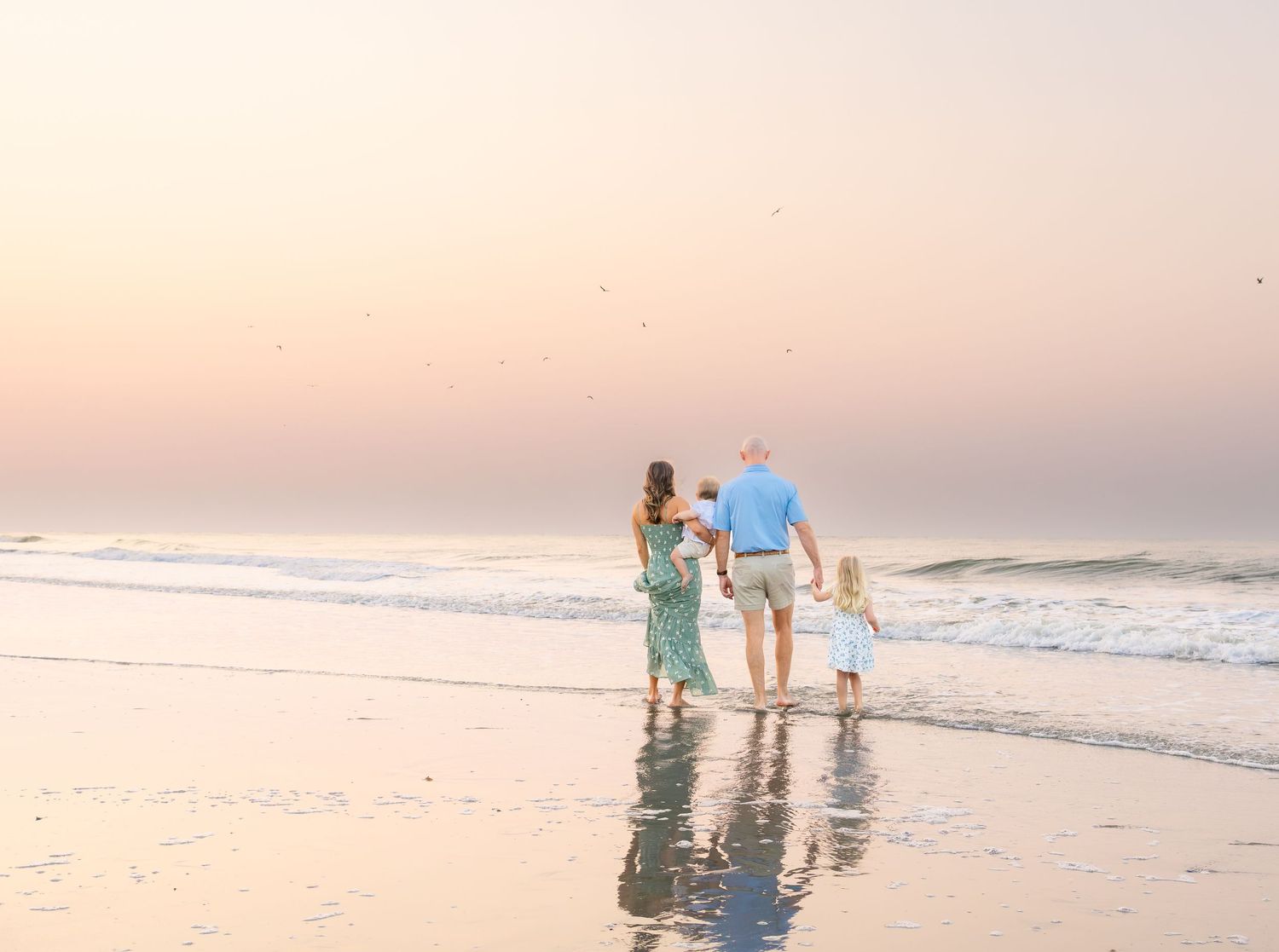 Family of three walking hand-in-hand along a serene beach at sunset with gentle waves.