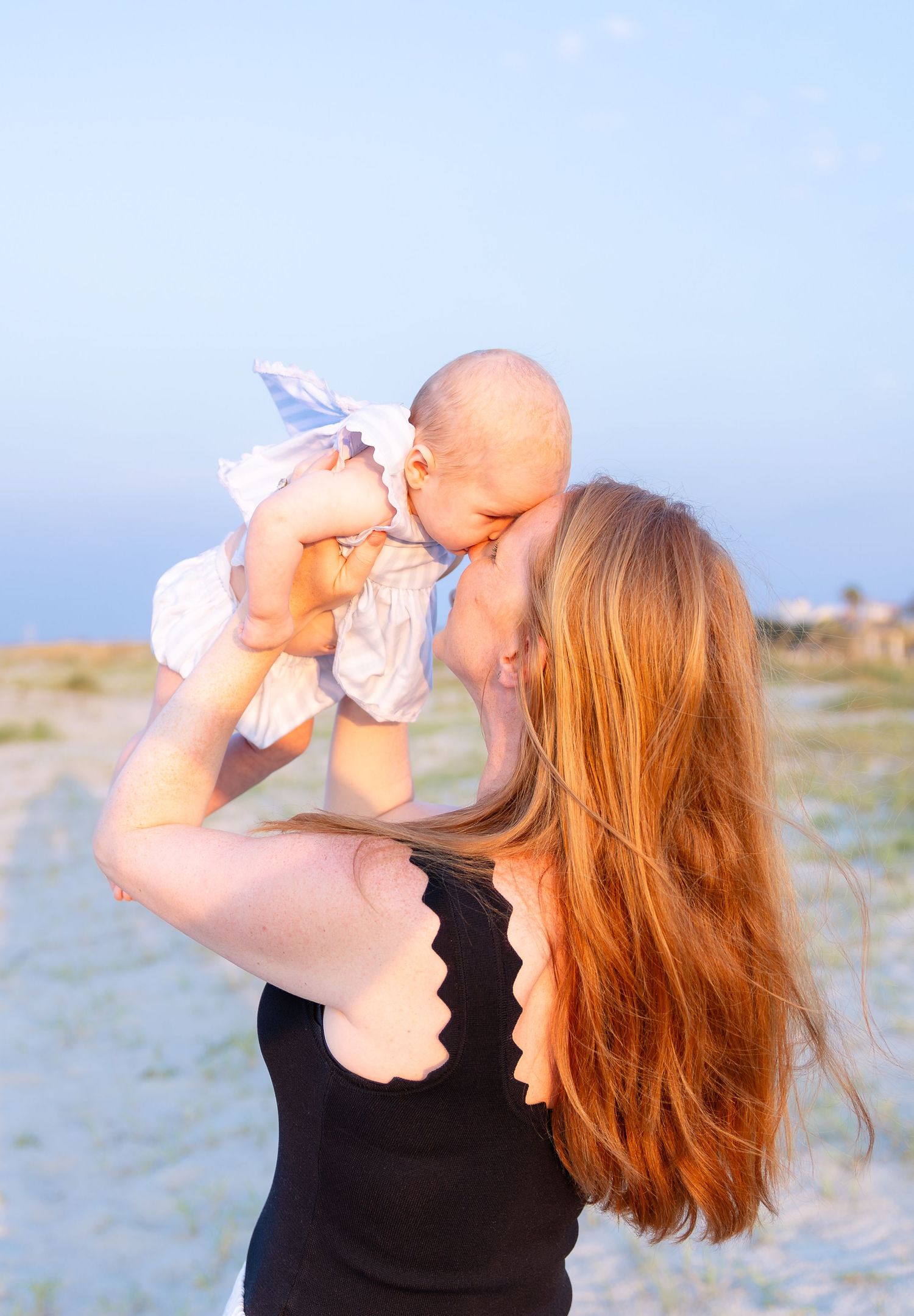A mother with long red hair holds her baby up to kiss at a sunny beach, wearing a black swimsuit.