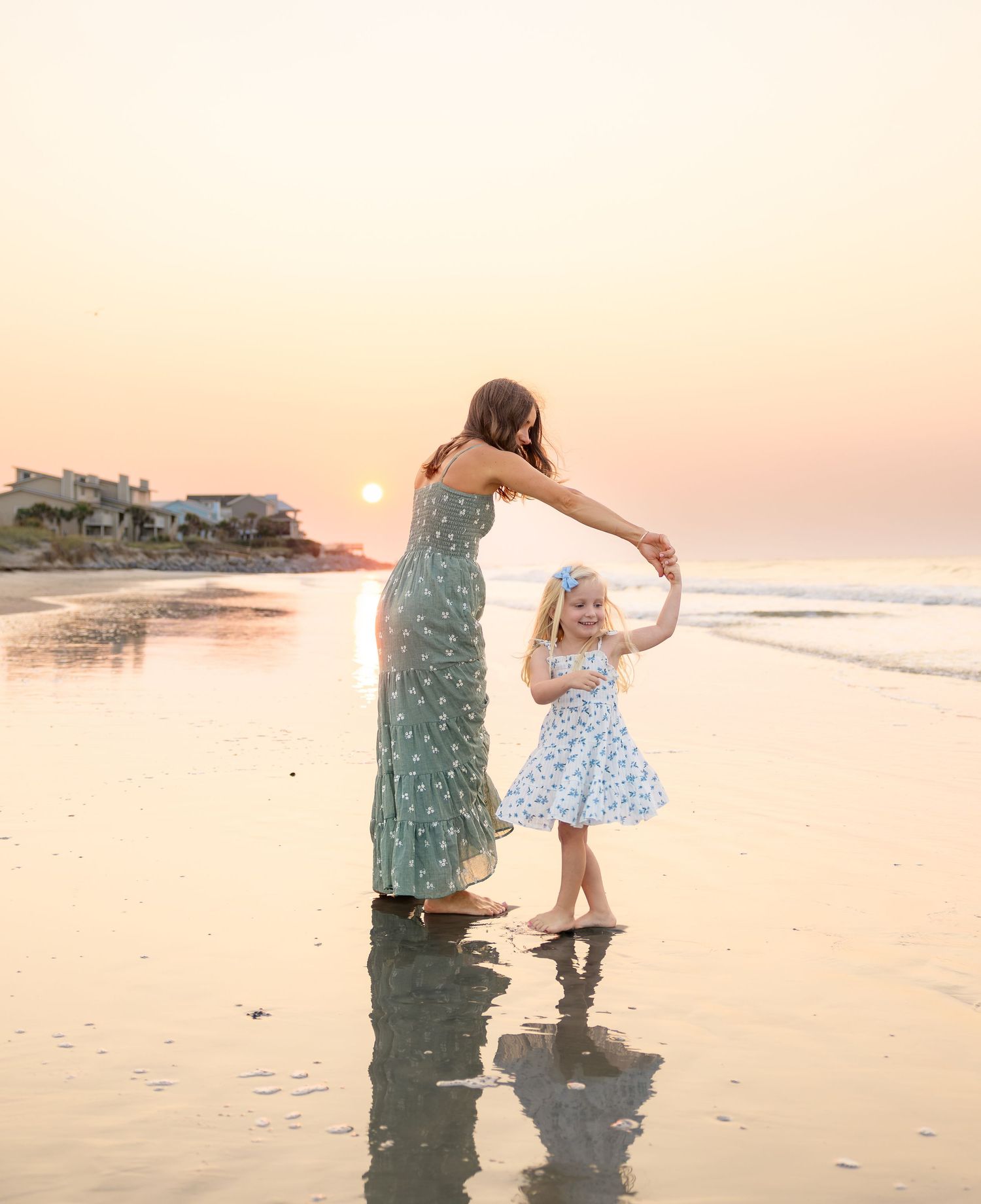 Mother and daughter dancing on a beach at golden sunset, wearing floral dresses, with reflections on wet sand.