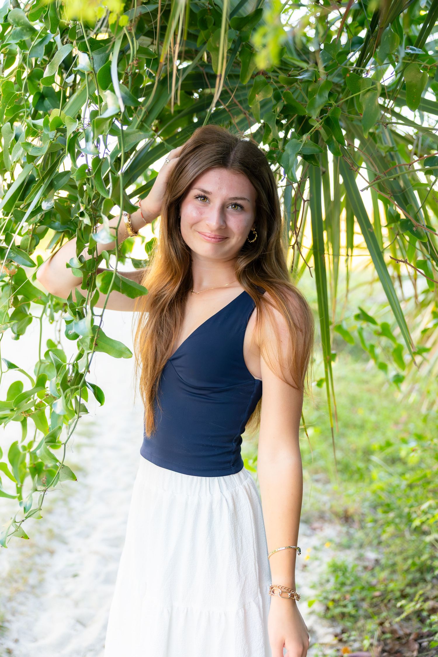 Smiling young woman with long auburn hair in a navy top and white skirt stands under green leafy branches on a sandy outdoor path.