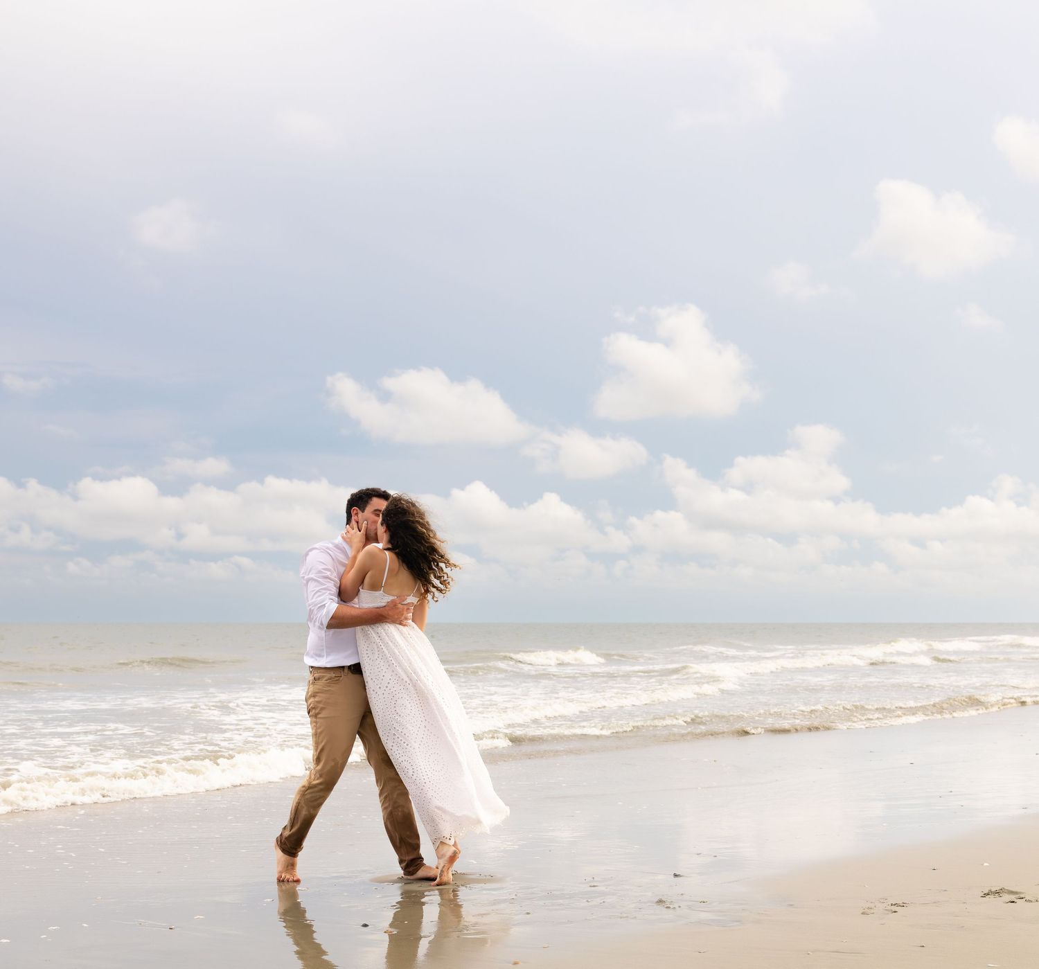 Romantic couple embracing and walking along a sandy beach shoreline with ocean waves and cloudy sky.