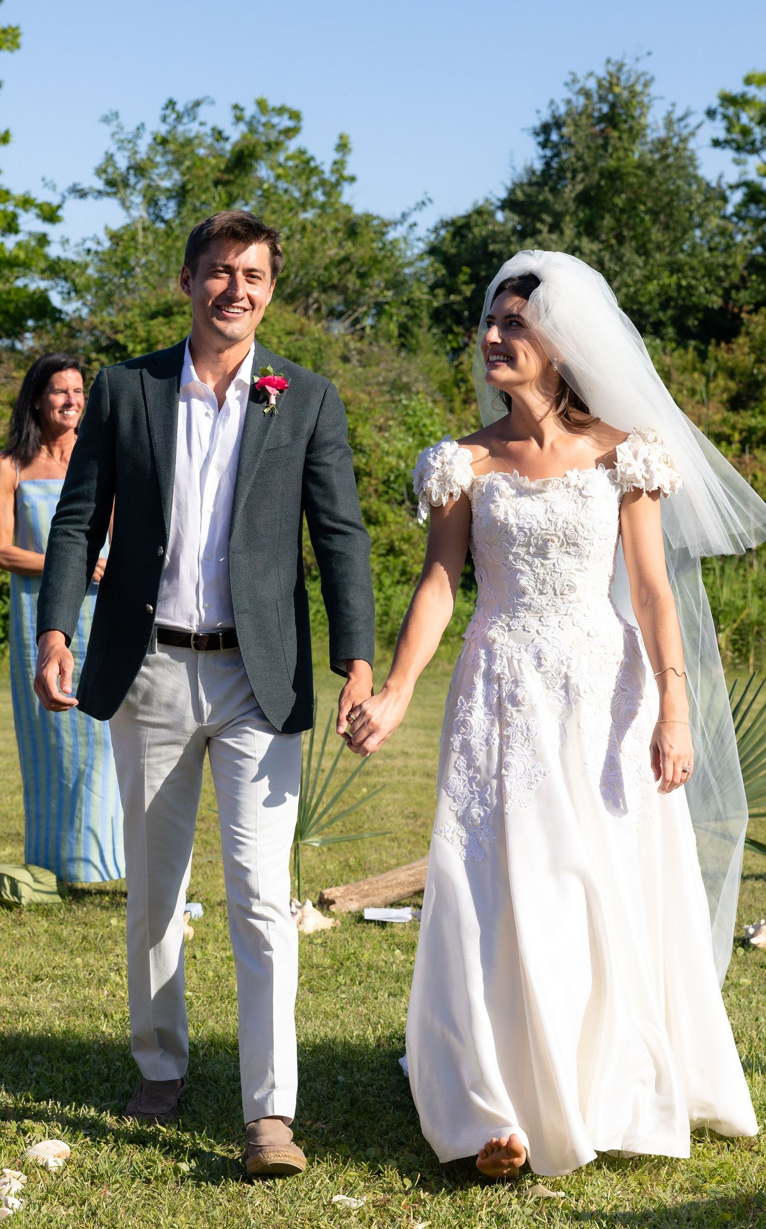 Smiling newlywed couple walks hand in hand outdoors after wedding ceremony, bride in lace gown with veil.