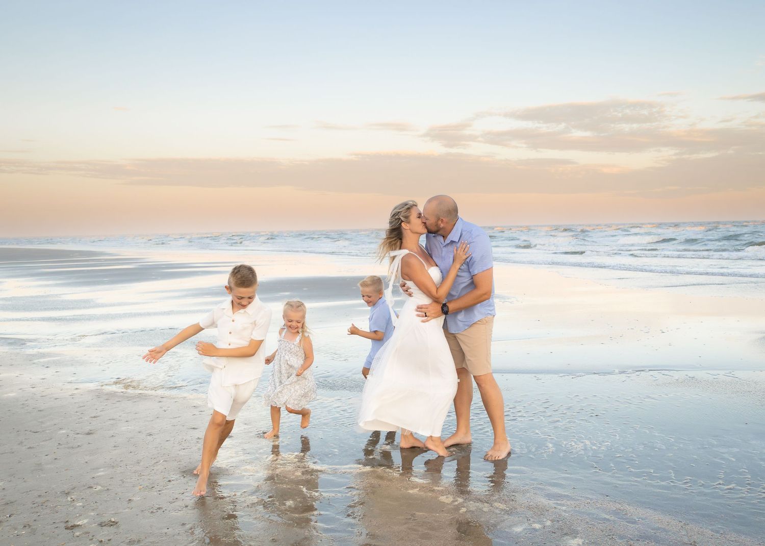 Family of five sharing a romantic kiss on a beach at sunset with children playing nearby.