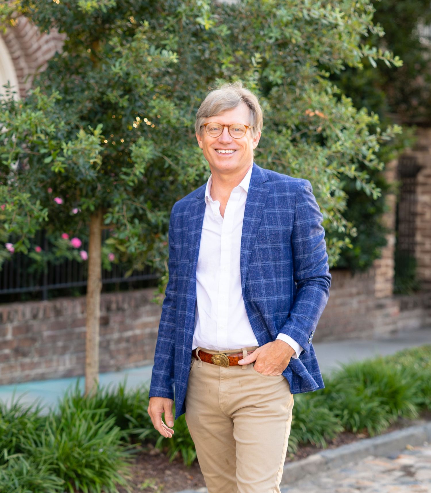 Smiling man in blue plaid blazer and khakis poses outdoors among lush green hedges and brick wall.