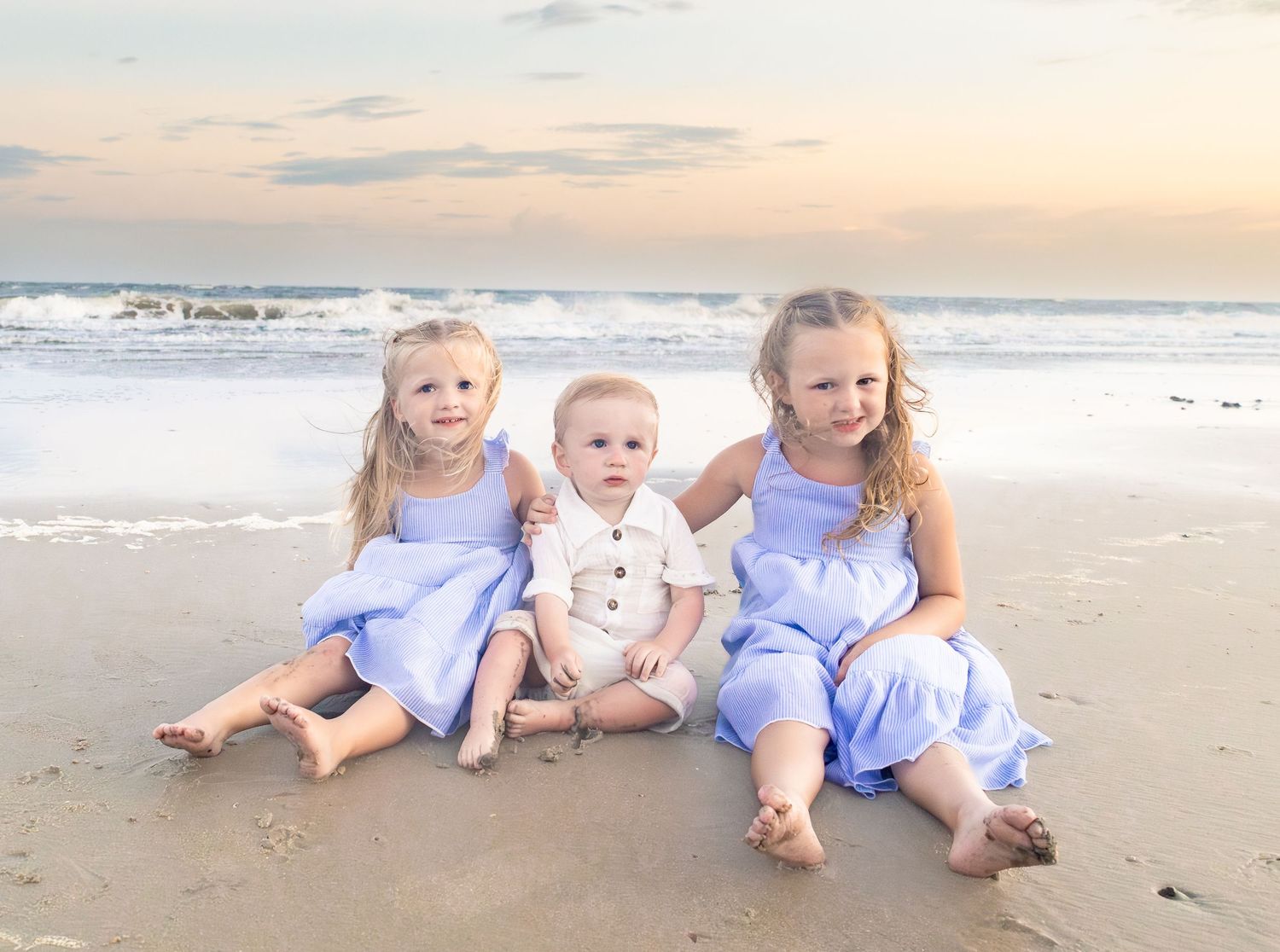 Three young children in matching blue outfits sitting together on a sandy beach at sunset.
