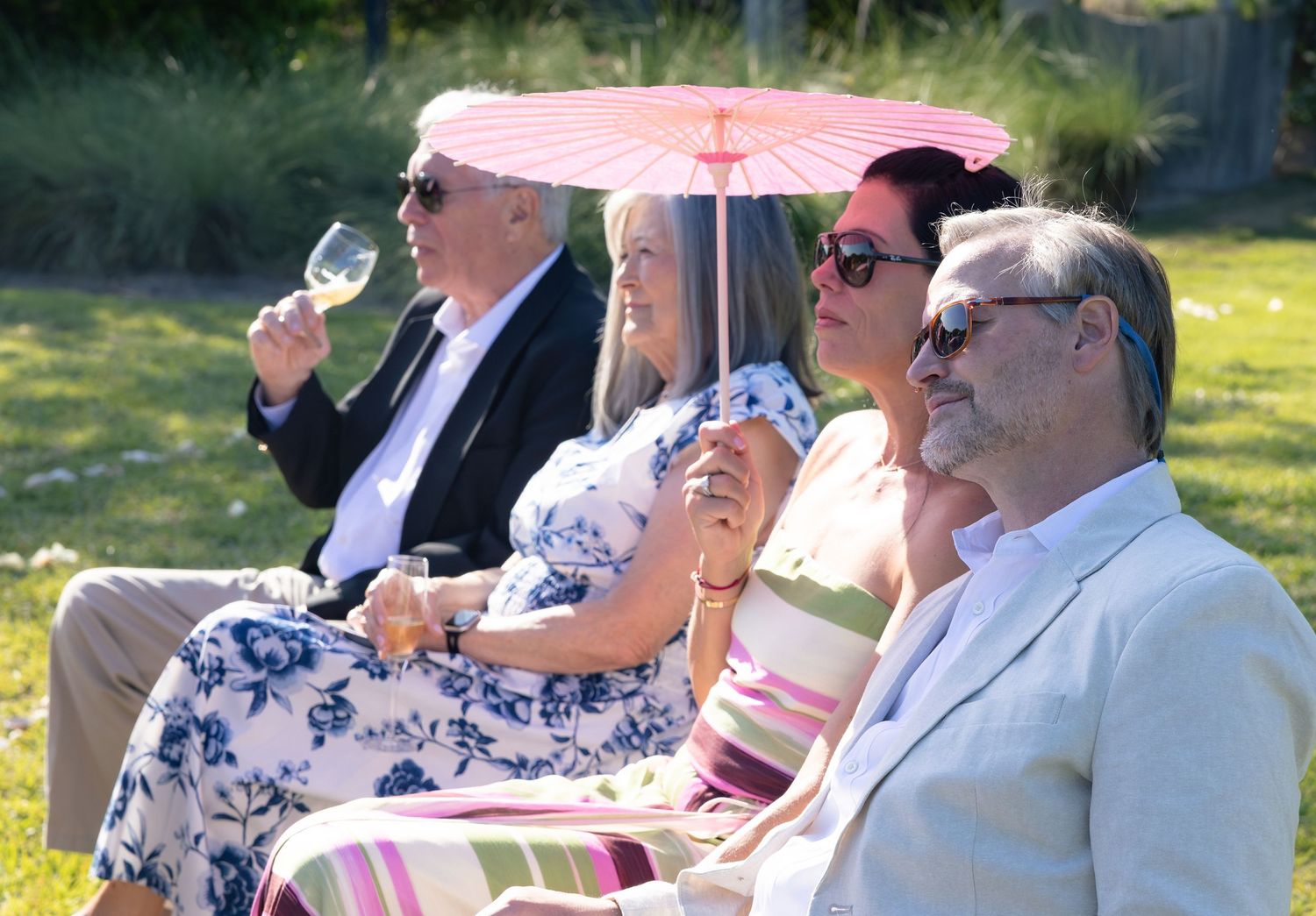 Group of adults relaxing on grass at outdoor garden party, one holding wine glass under pink parasol.
