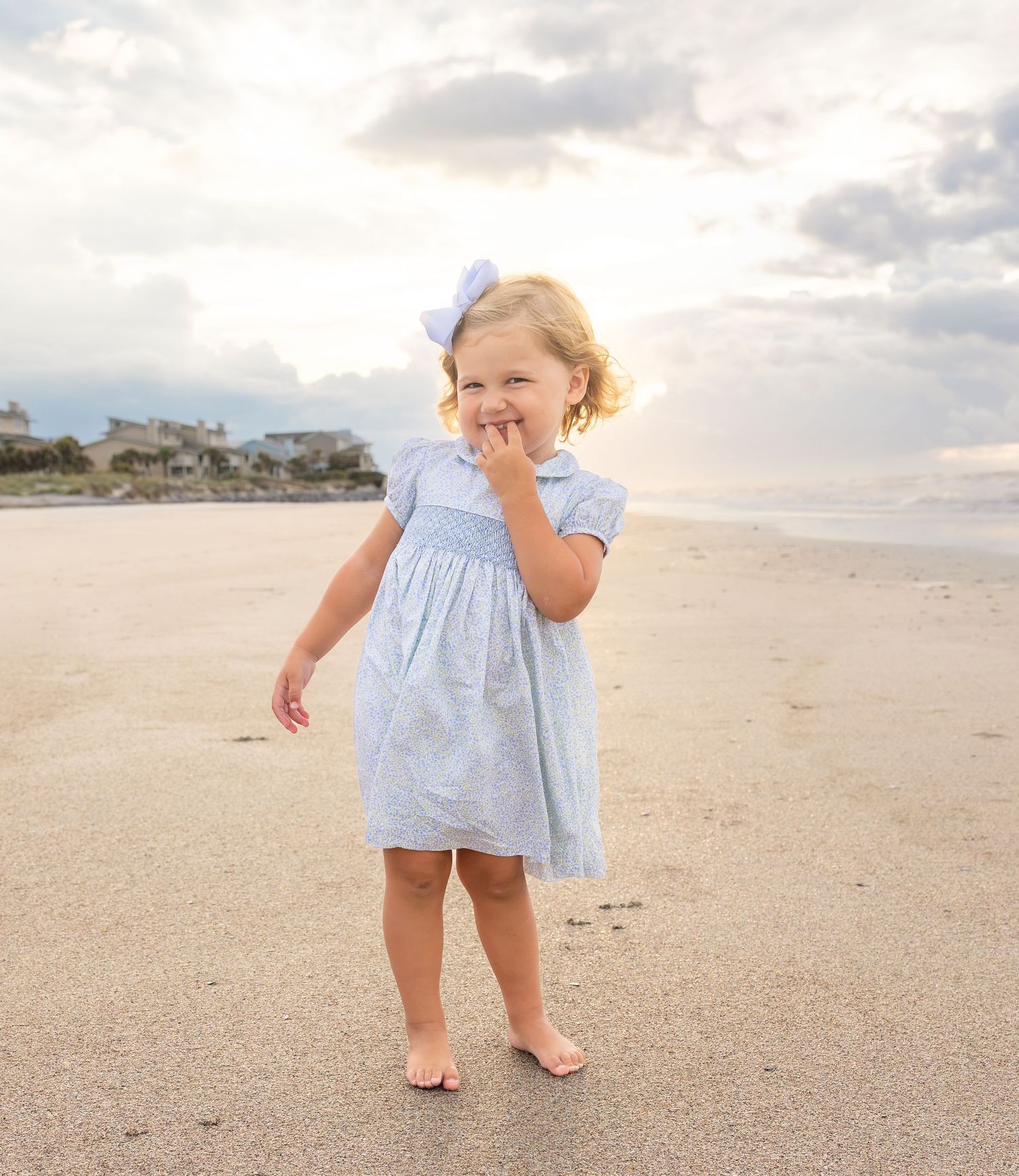 A young toddler girl in a light blue dress and bow stands barefoot on a sandy beach with cloudy skies.