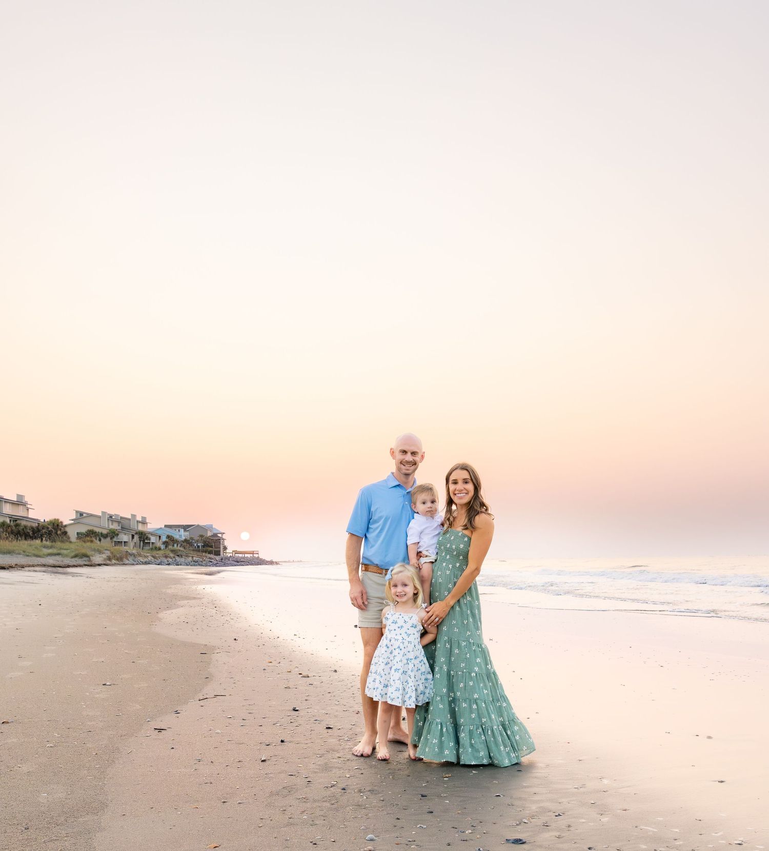 Family of four posing on a sandy beach at sunset, wearing pastel outfits with ocean waves in the background.