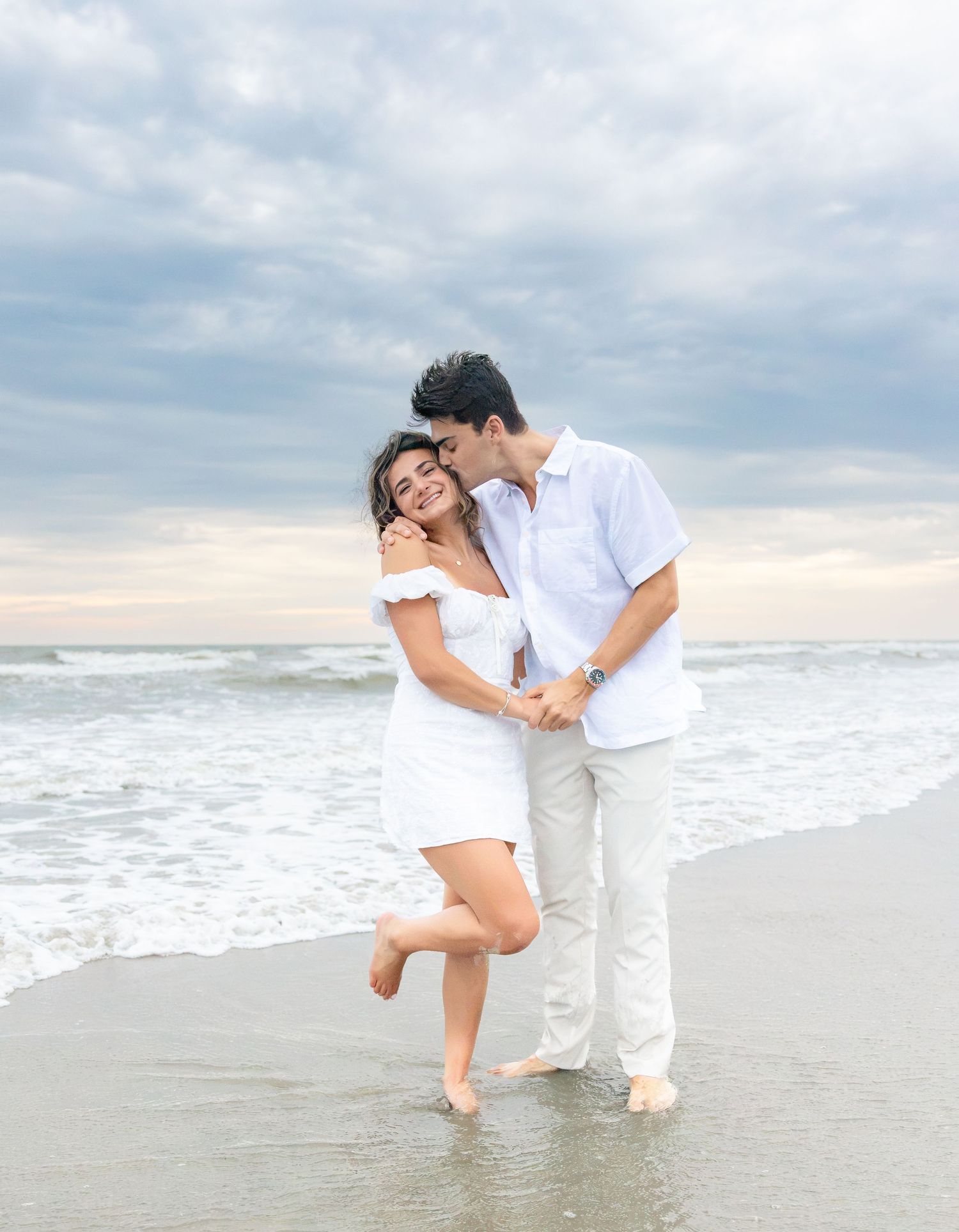 A couple in white outfits embraces romantically on a beach, with ocean waves and cloudy sky behind them.