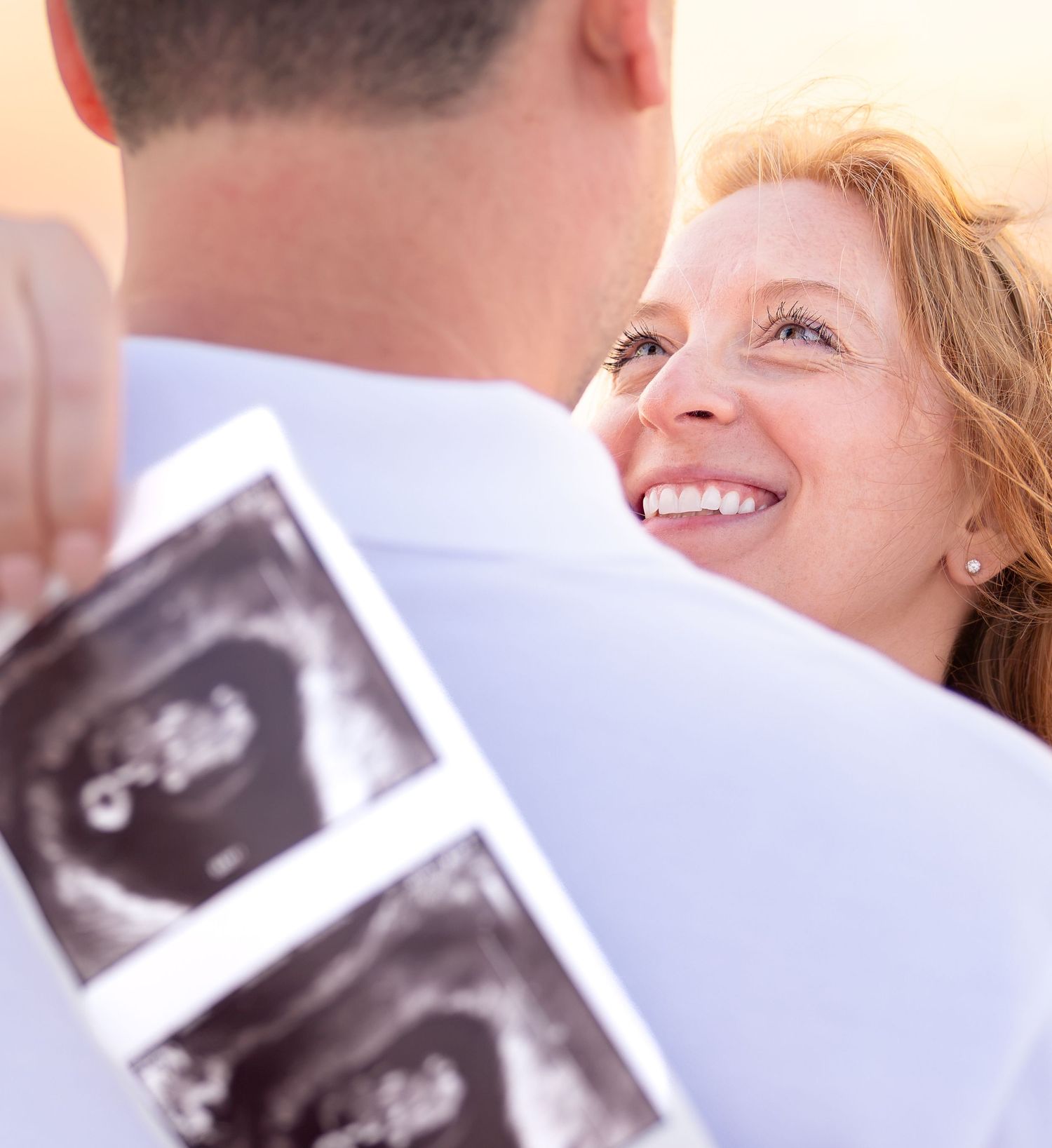 Happy couple embracing outdoors, woman smiling while man holds ultrasound photos, announcing pregnancy.