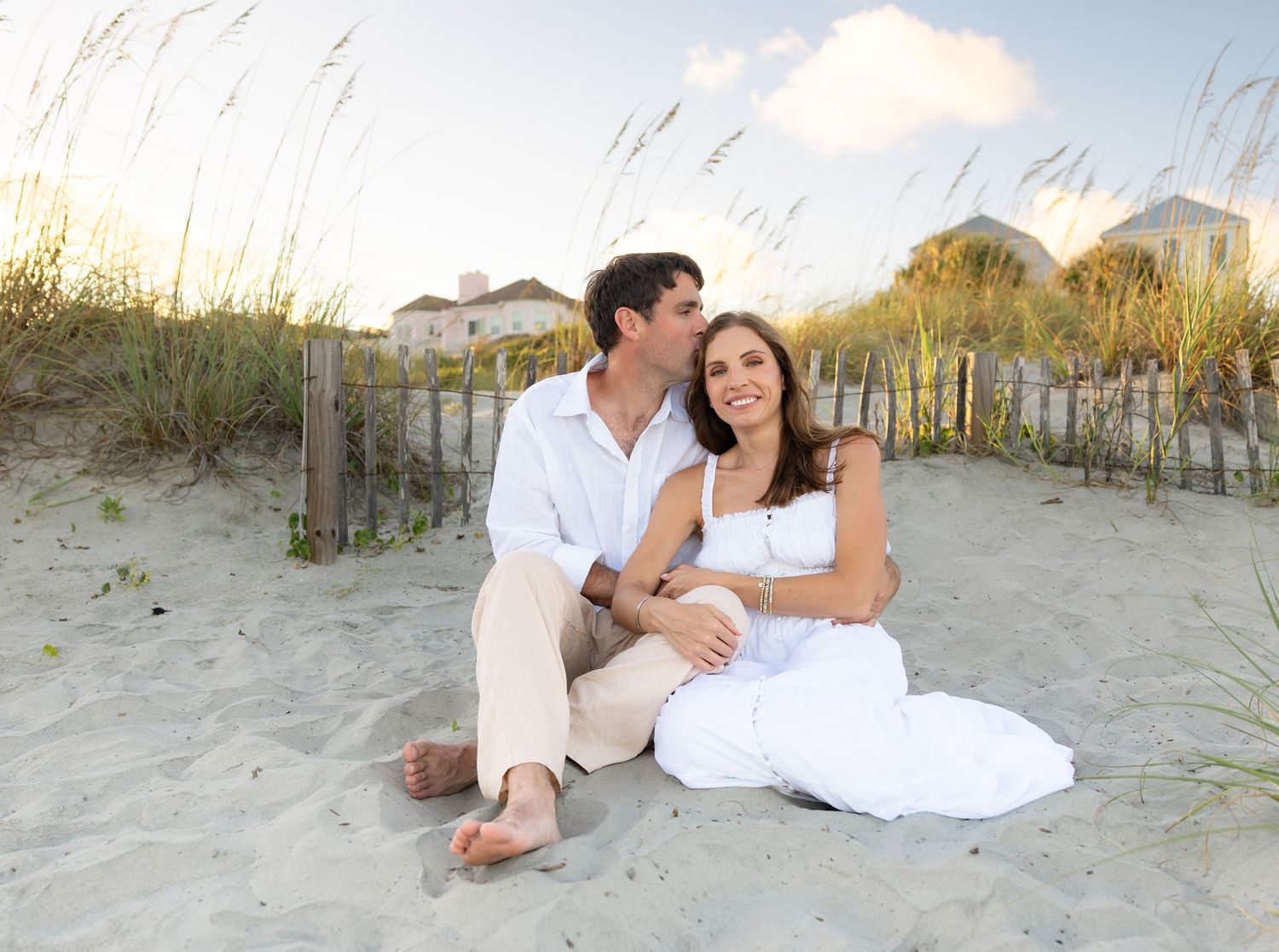 Romantic couple sitting on beach sand dunes in white outfits, woman looking up at man with sea oats behind them.