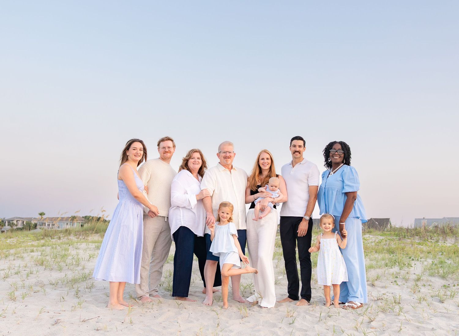 Large family group portrait on a sandy beach at sunset, wearing coordinated pastel outfits with children and adults smiling.