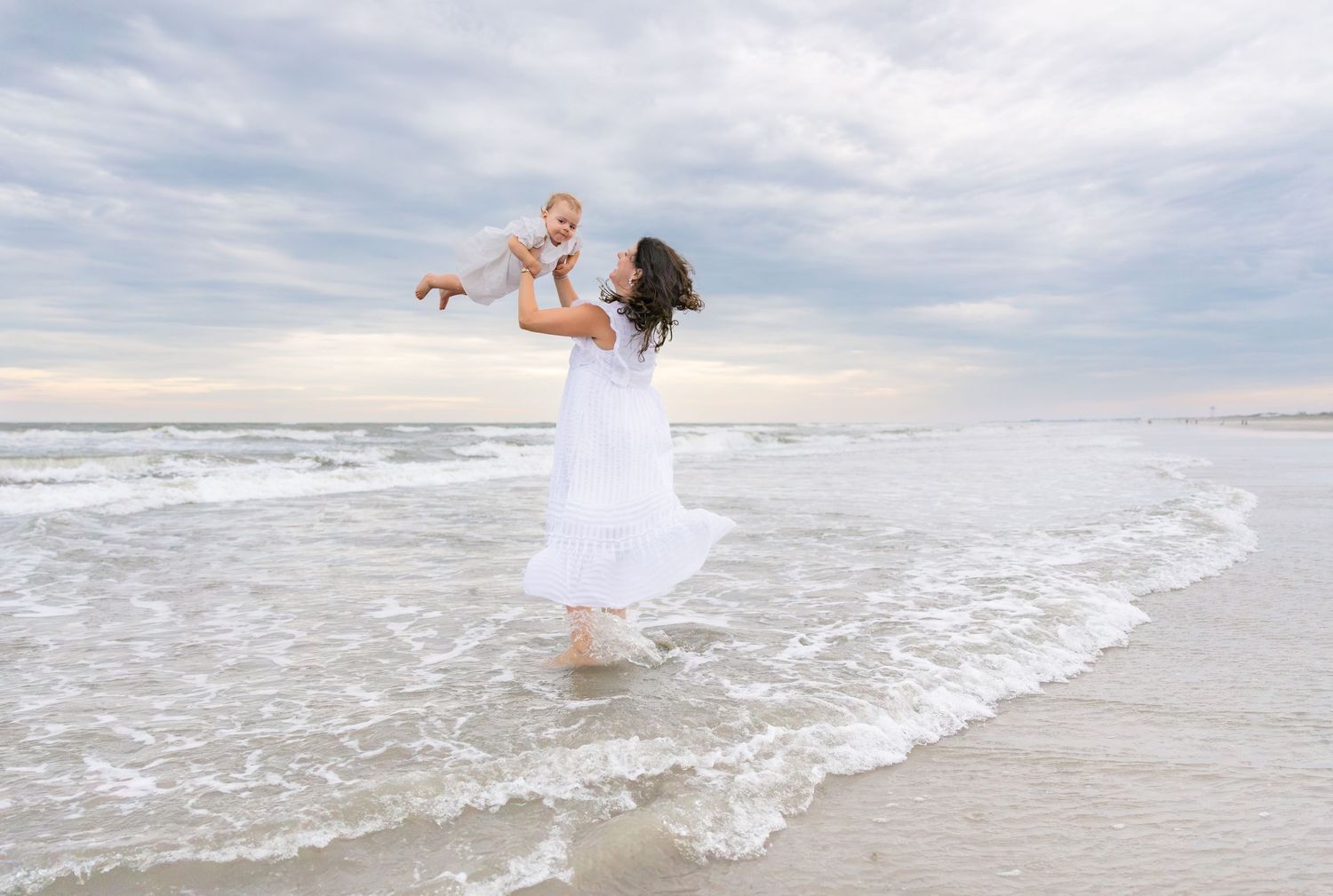 Mother in white dress lifts baby joyfully at ocean shoreline on cloudy day.