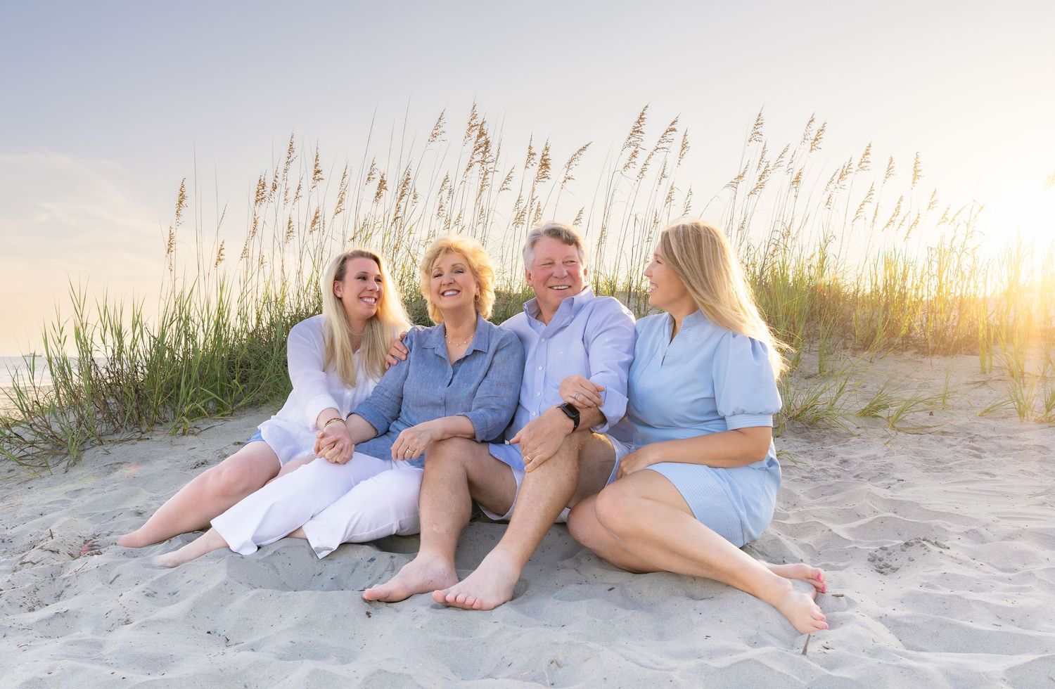 Family of four smiling together on a sandy beach at sunset with sea oats in the background.