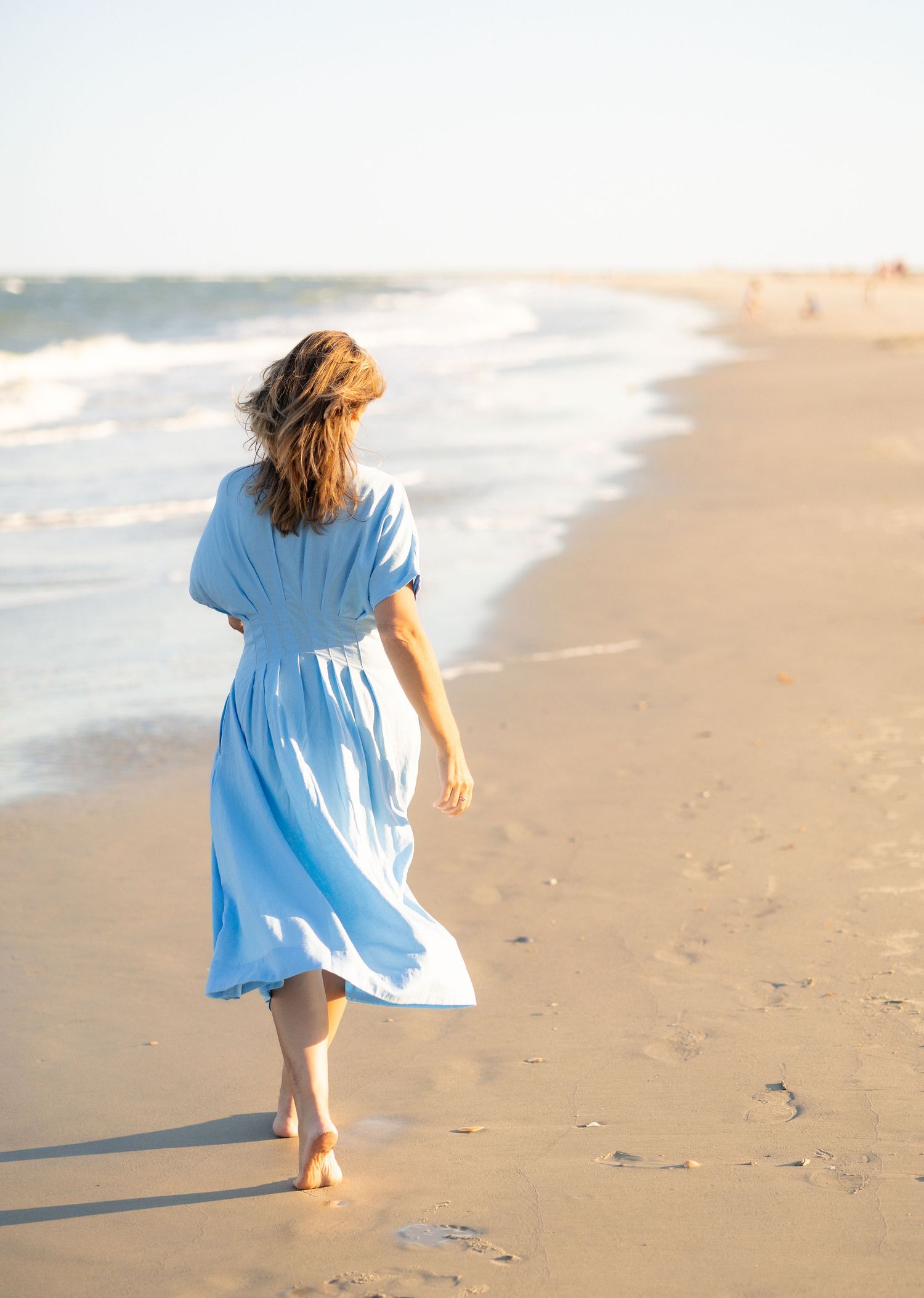 Woman in blue midi dress walking barefoot on sandy beach with ocean waves in background.