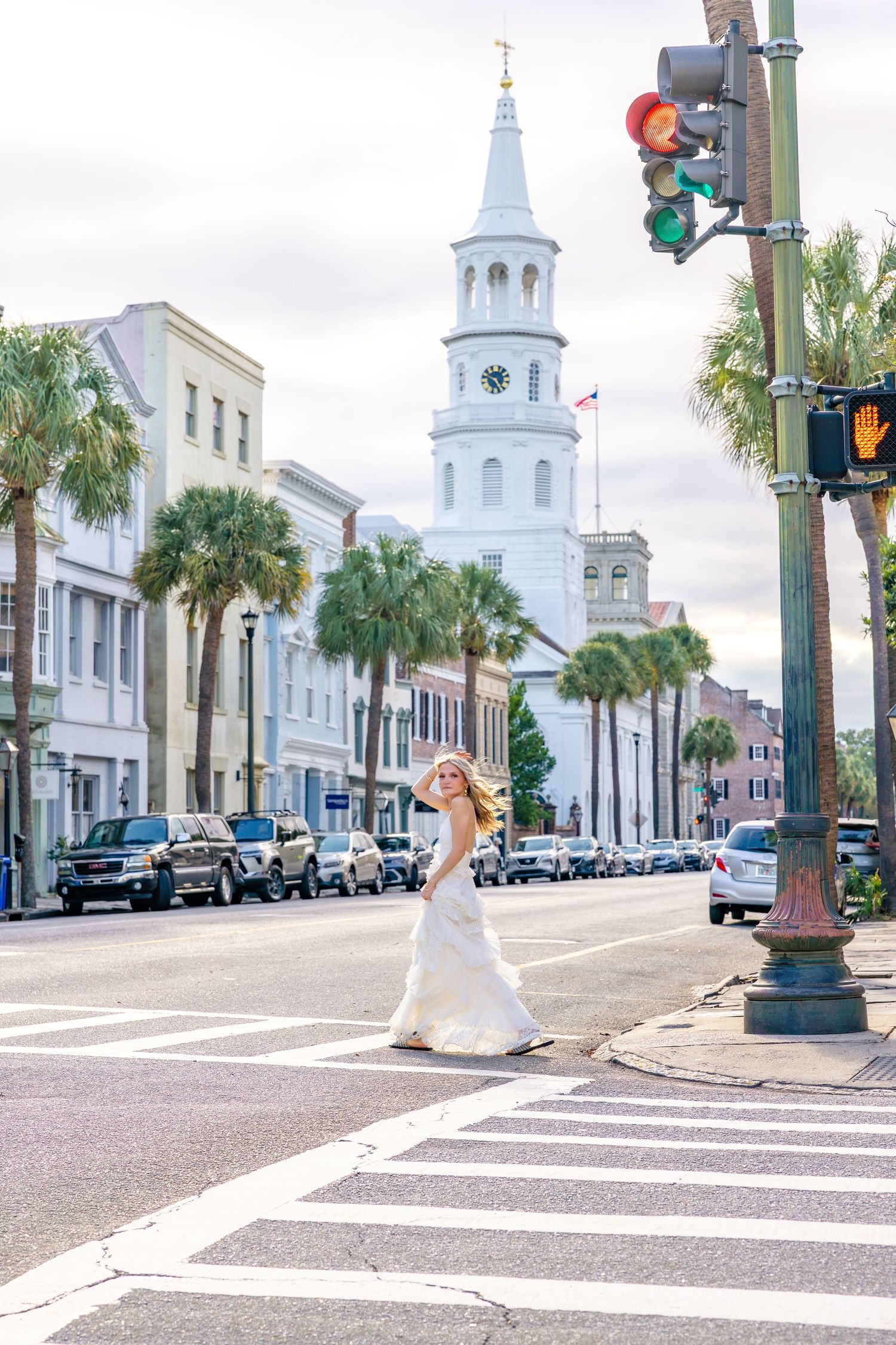 Woman in a flowing white gown crosses a downtown crosswalk with palm trees and a white church steeple in Charleston, South Carolina.