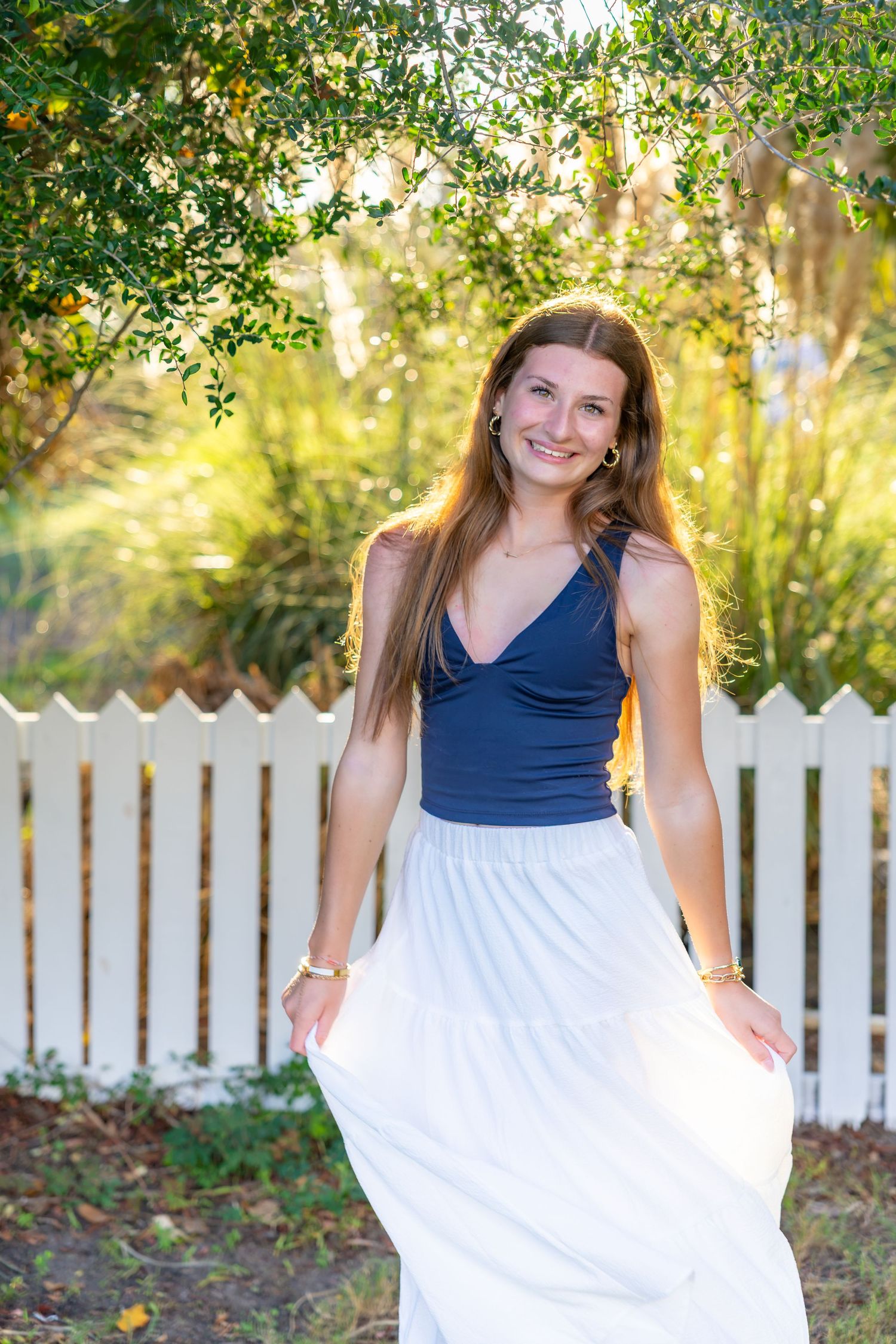 Smiling young woman with long hair in a navy tank top and flowing white skirt poses outdoors near a white picket fence and green foliage.