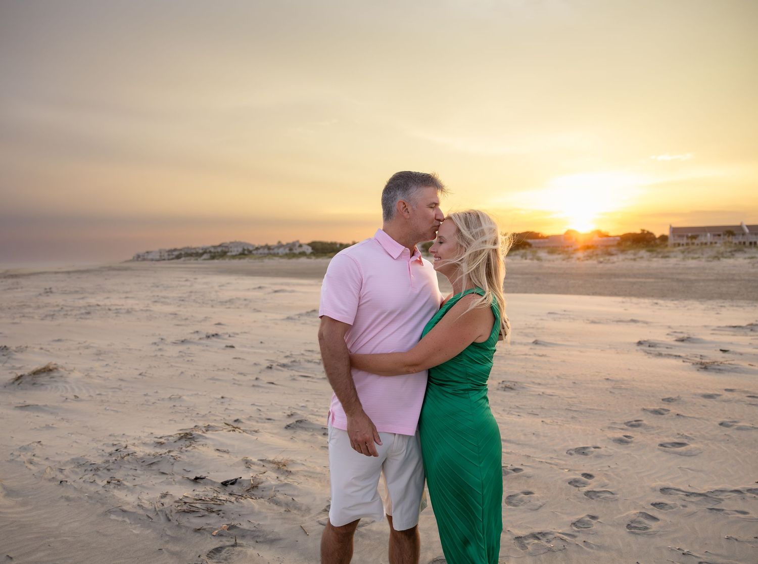 Couple embracing on a sandy beach at sunset, the man in a pink polo kissing the woman in a green dress on the forehead.