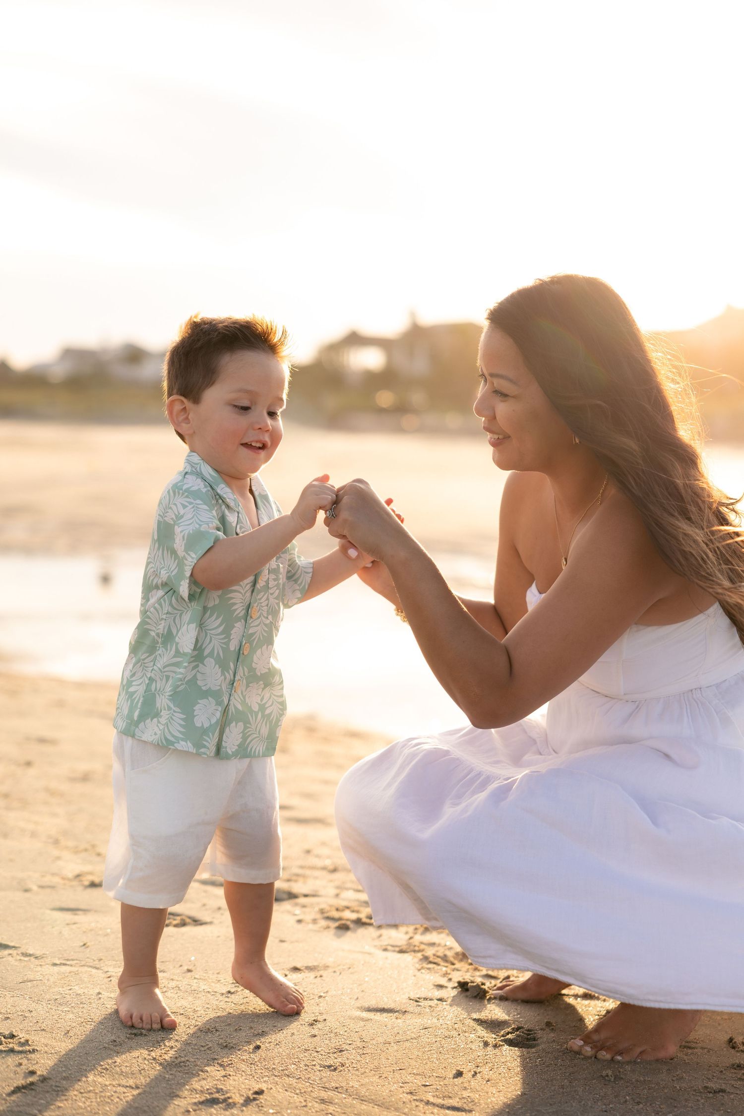 Mother and toddler son holding hands on a sandy beach at golden sunset, boy wearing Hawaiian shirt.