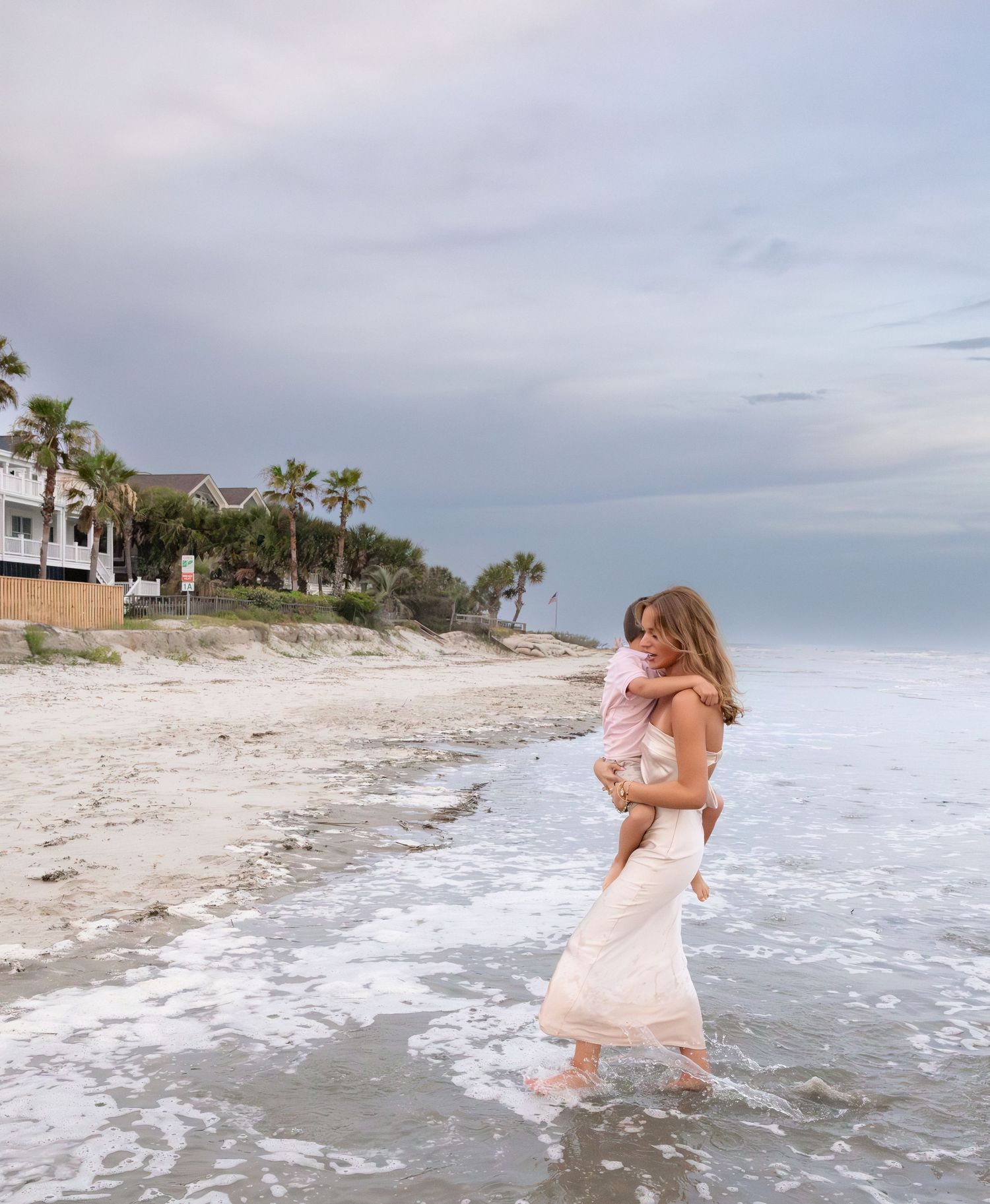 A woman in a white dress carries a toddler while wading along a sandy beach with palm trees and houses in the background.