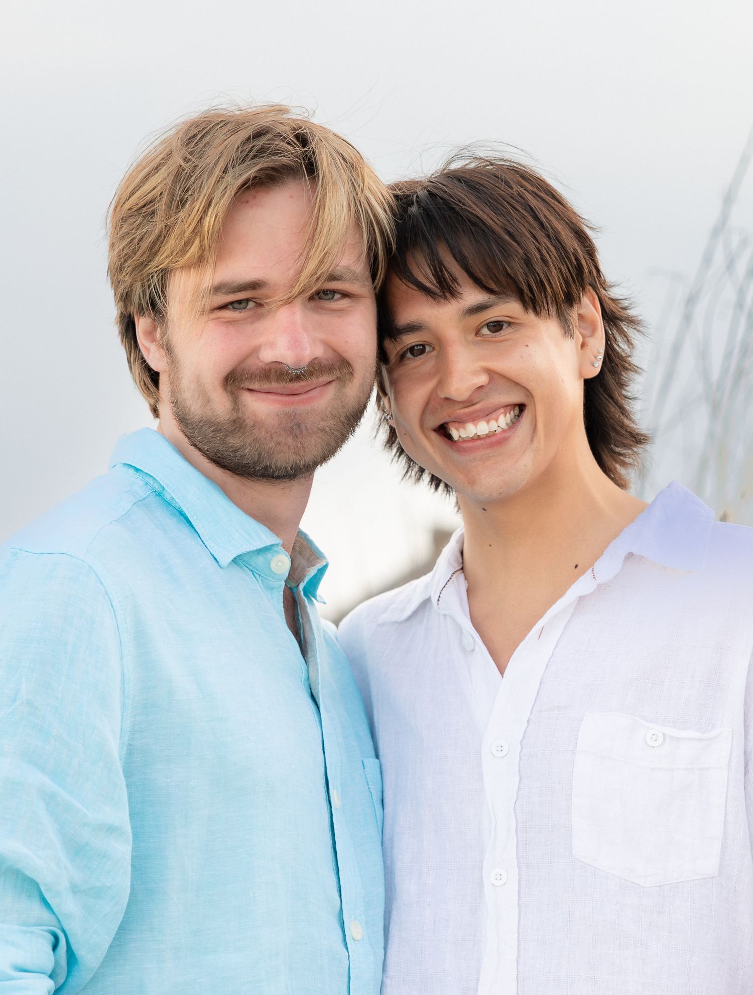 Two smiling young men in light blue and white shirts posing together outdoors in a bright, sunny setting.