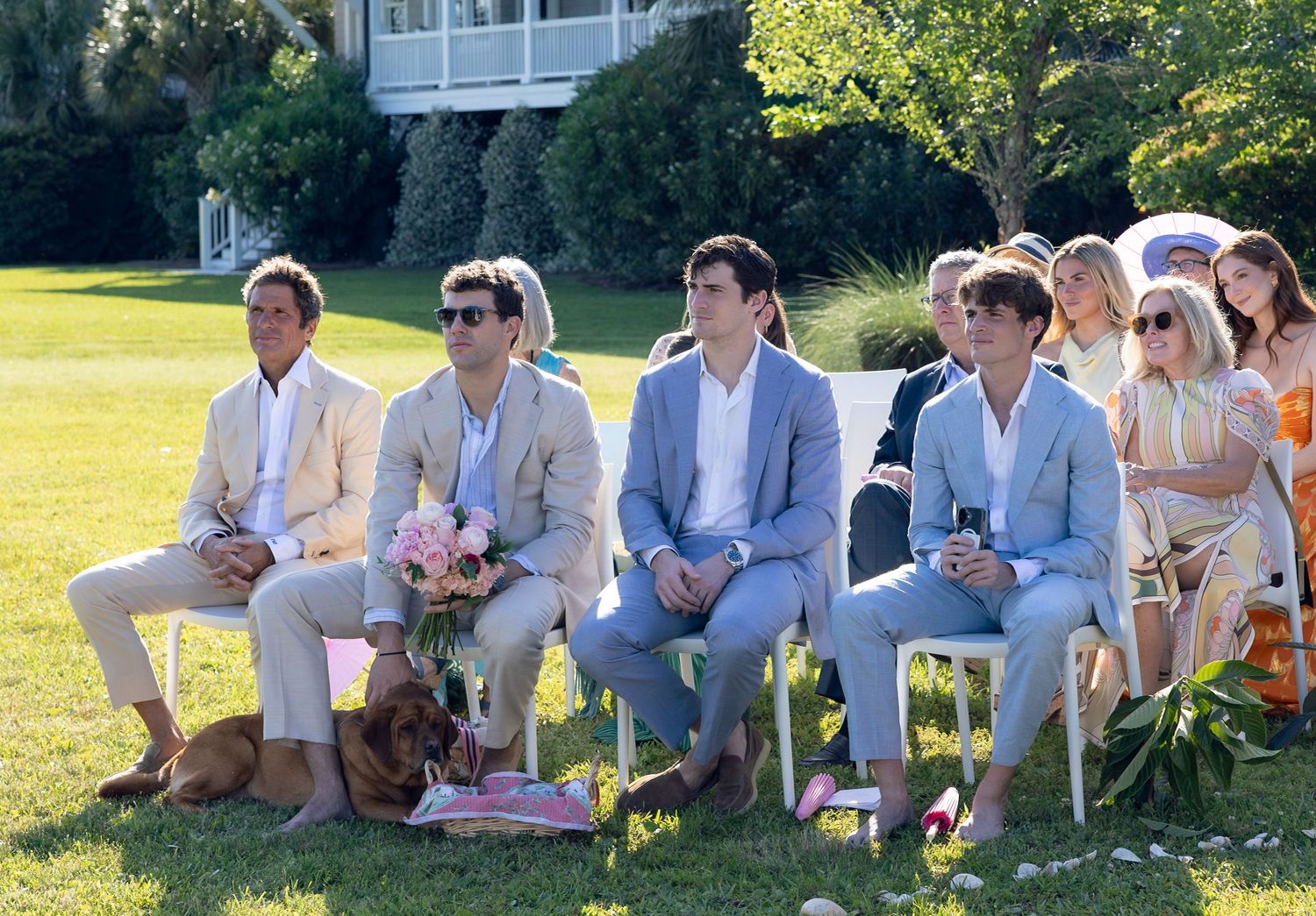 Guests seated in white chairs at an outdoor wedding ceremony on a green lawn with a dachshund dog.