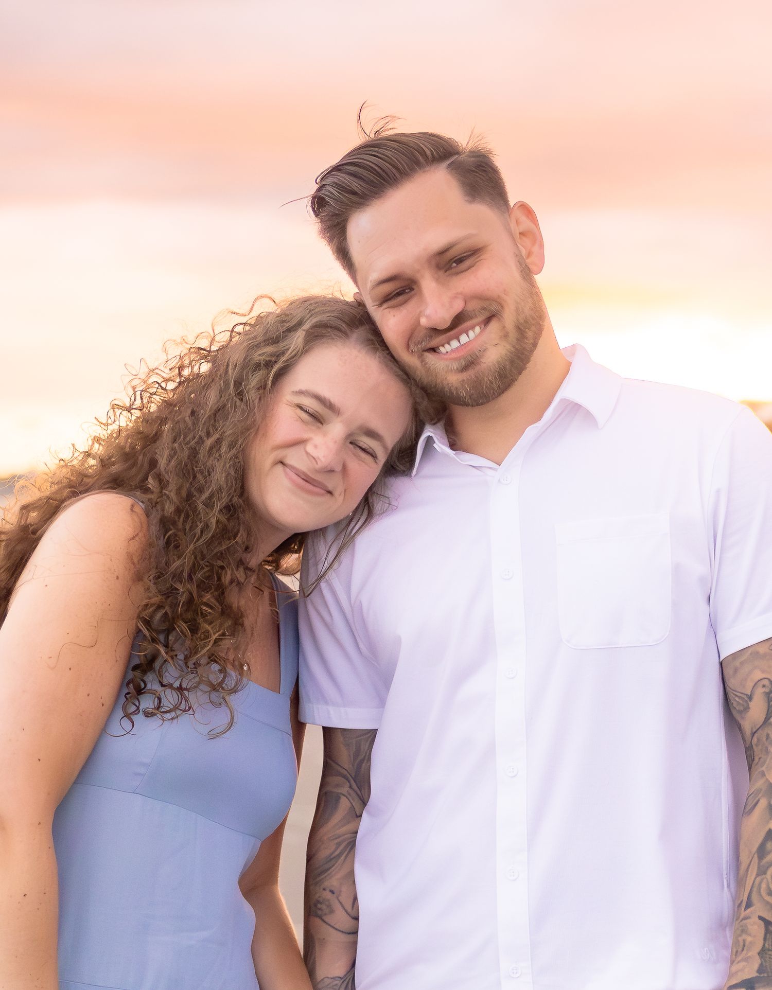 Smiling engaged couple poses together at beach sunset, woman resting head on mans shoulder.