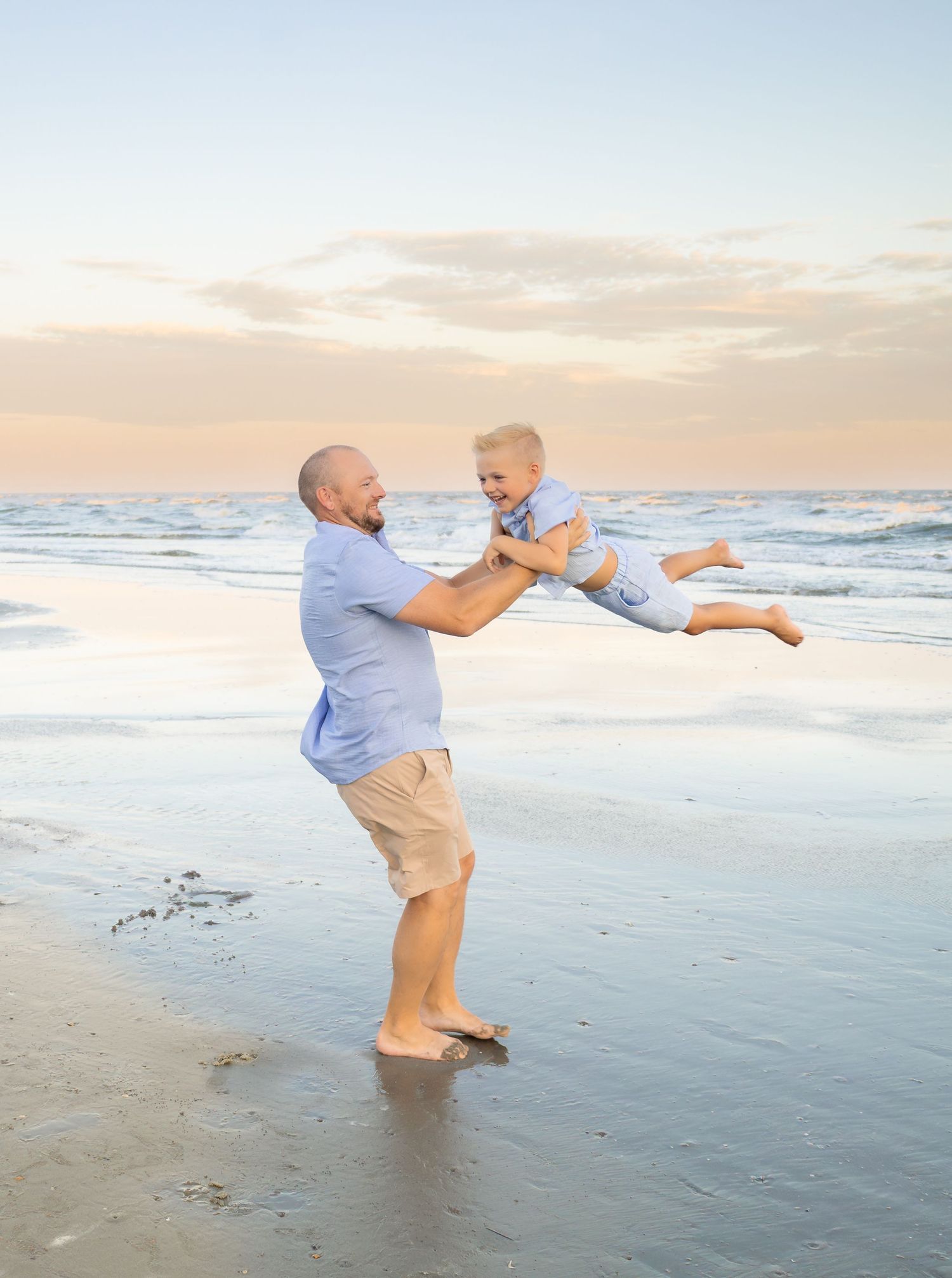 A father joyfully lifts his toddler in the air on a sandy beach at sunset, both wearing light blue outfits.