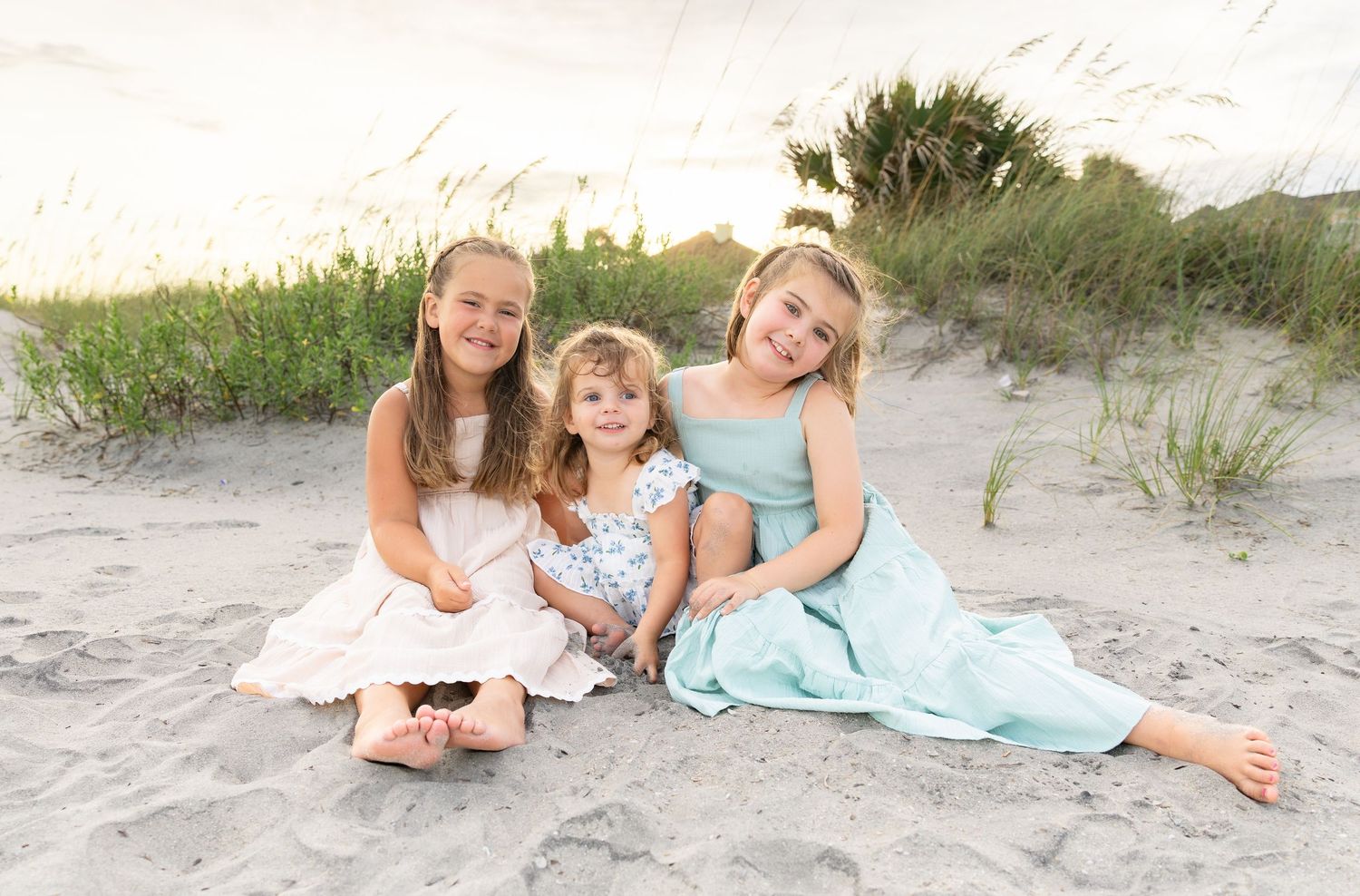 Three young girls in white and blue dresses sitting together on a sandy beach with dunes and grass in the background.