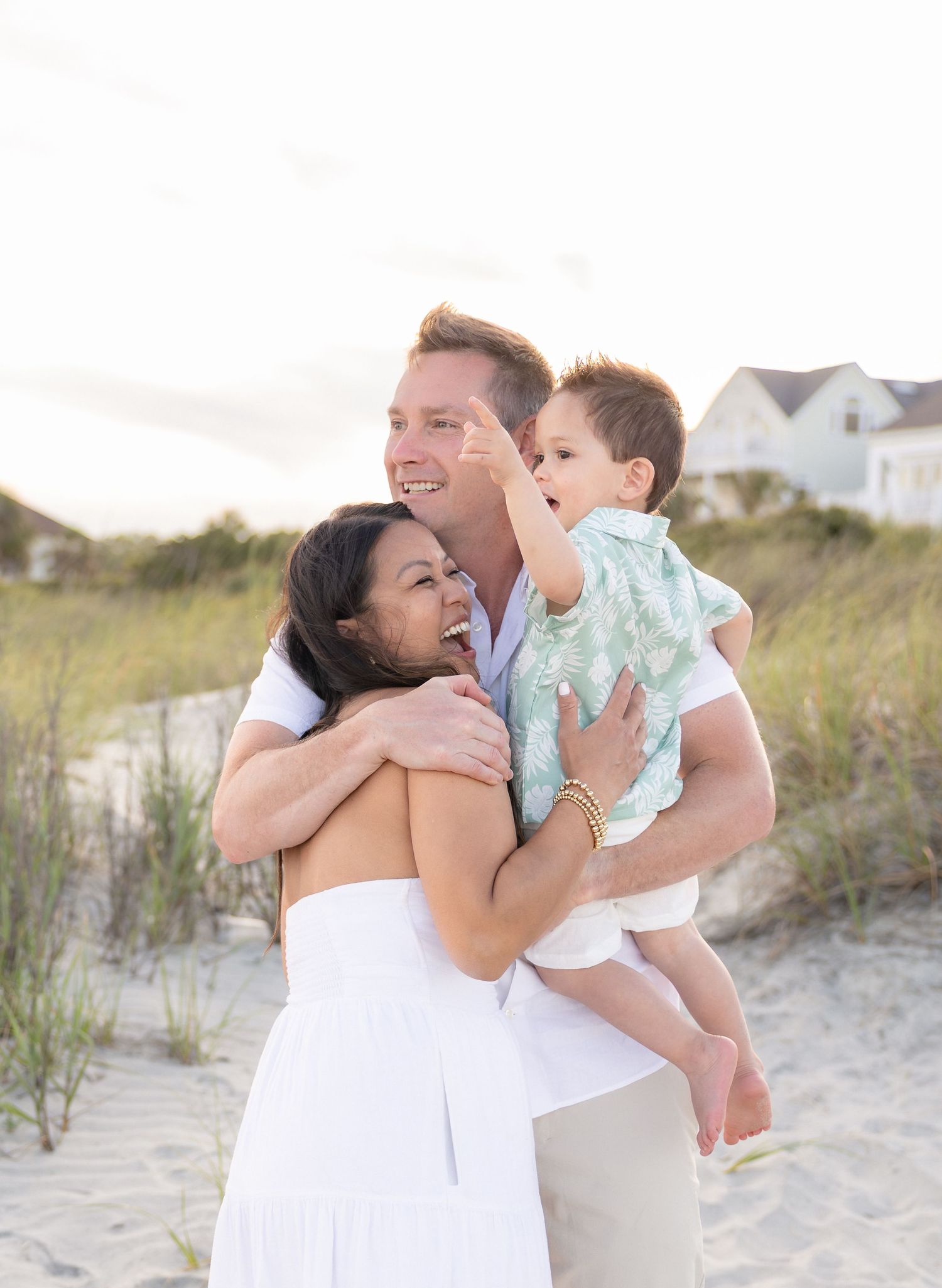 Happy family of three enjoying a beach photoshoot on sandy dunes, parents smiling as dad holds toddler in mint outfit.