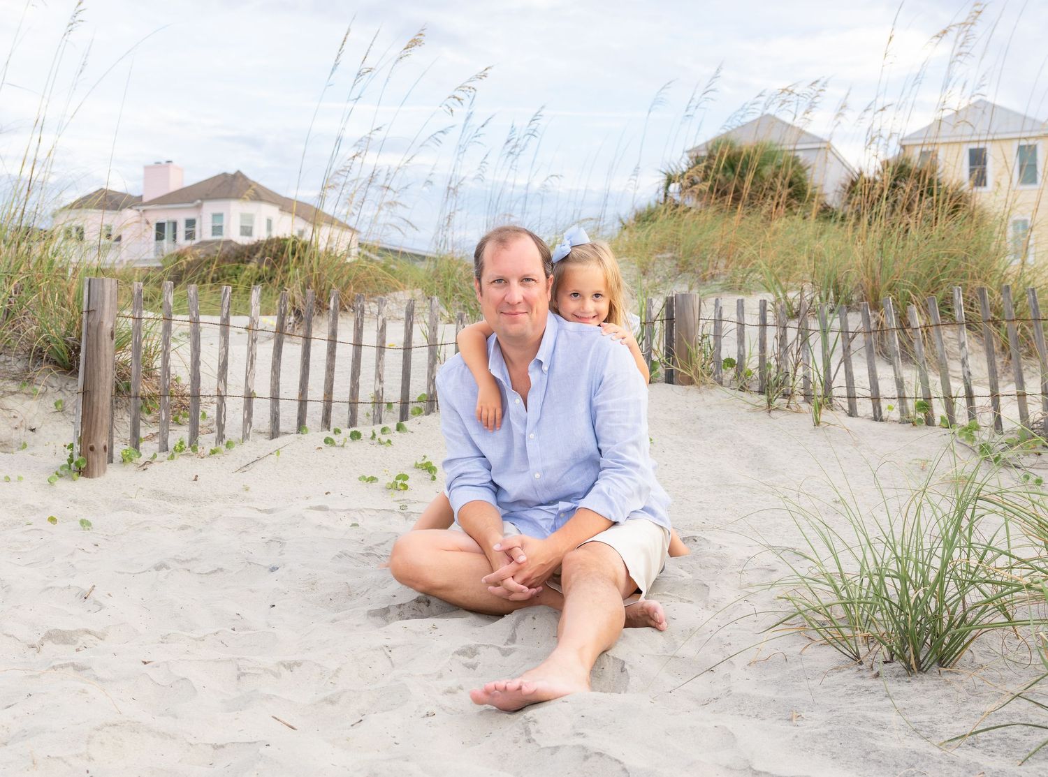 A man and young girl sit together on sandy beach dunes with sea grass and a wooden fence behind them.