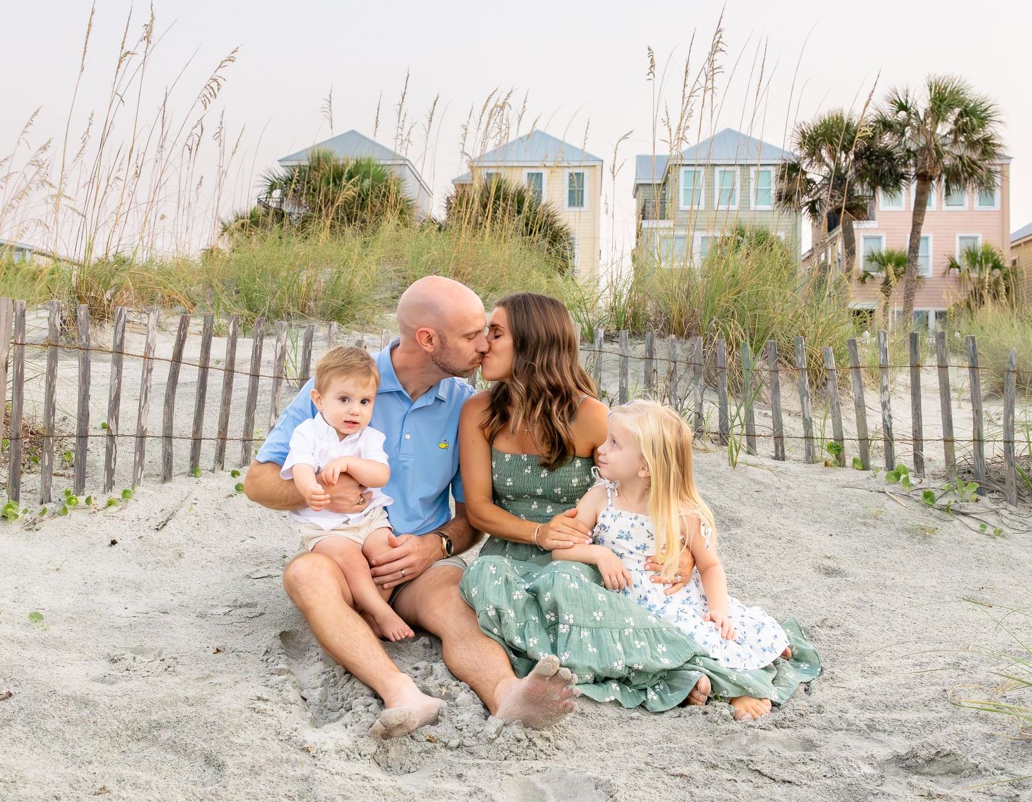 Family of four sitting on beach sand, parents kissing while holding toddler and young girl in matching outfits.