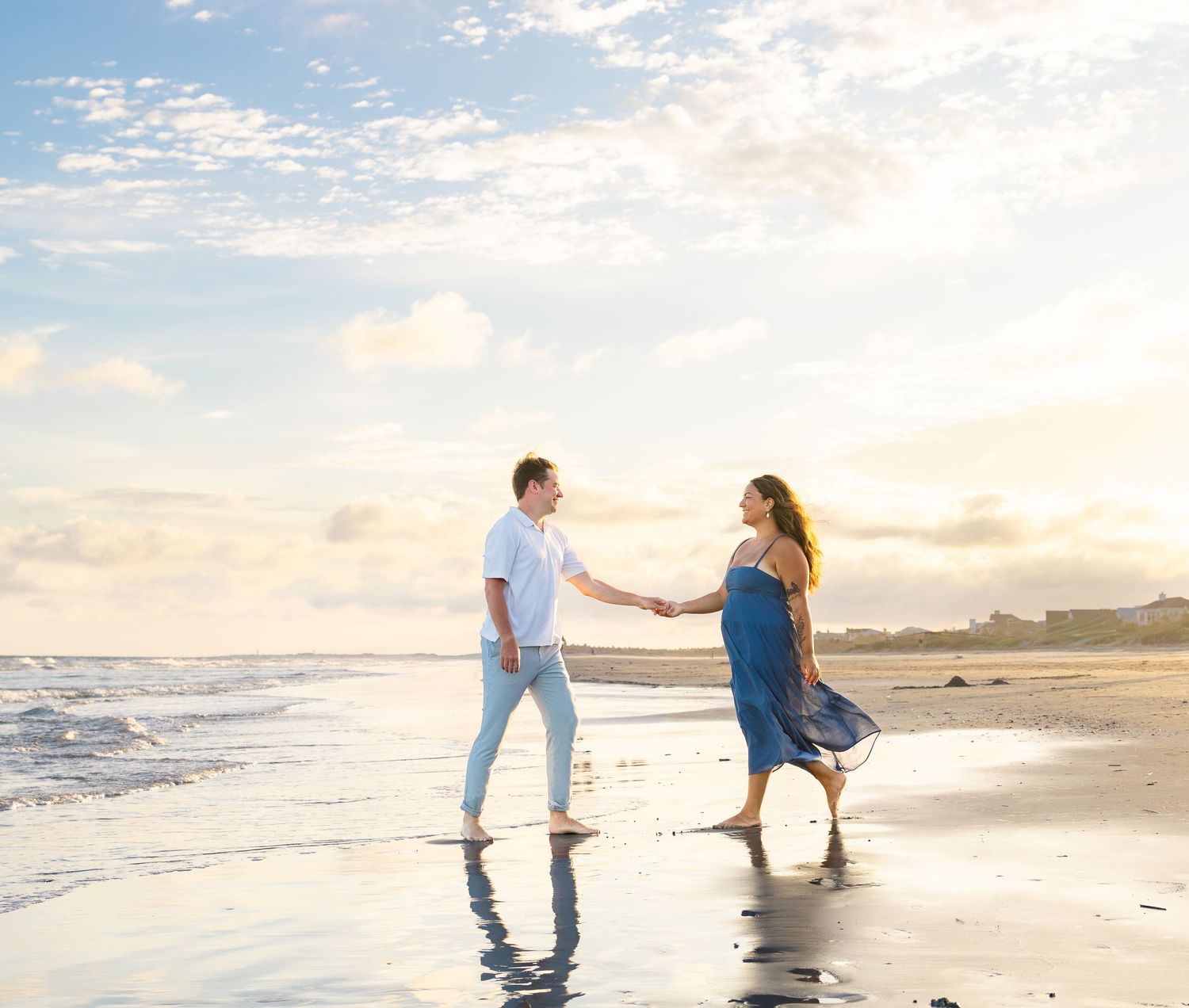 A couple holds hands and dances barefoot on a sandy beach at sunset with gentle waves and a colorful sky.