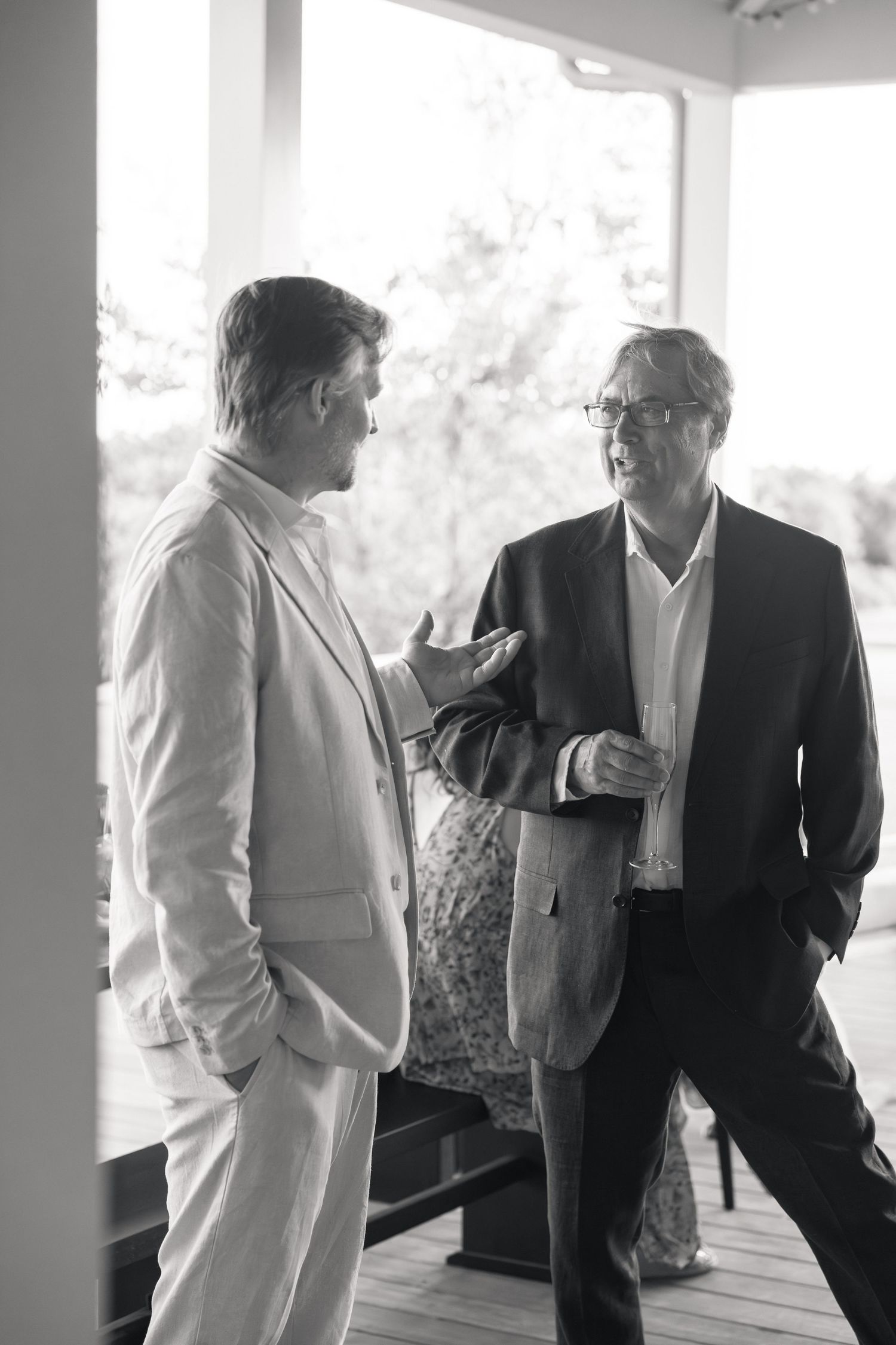Two men in formal attire having a conversation on a covered porch in black and white wedding photography.