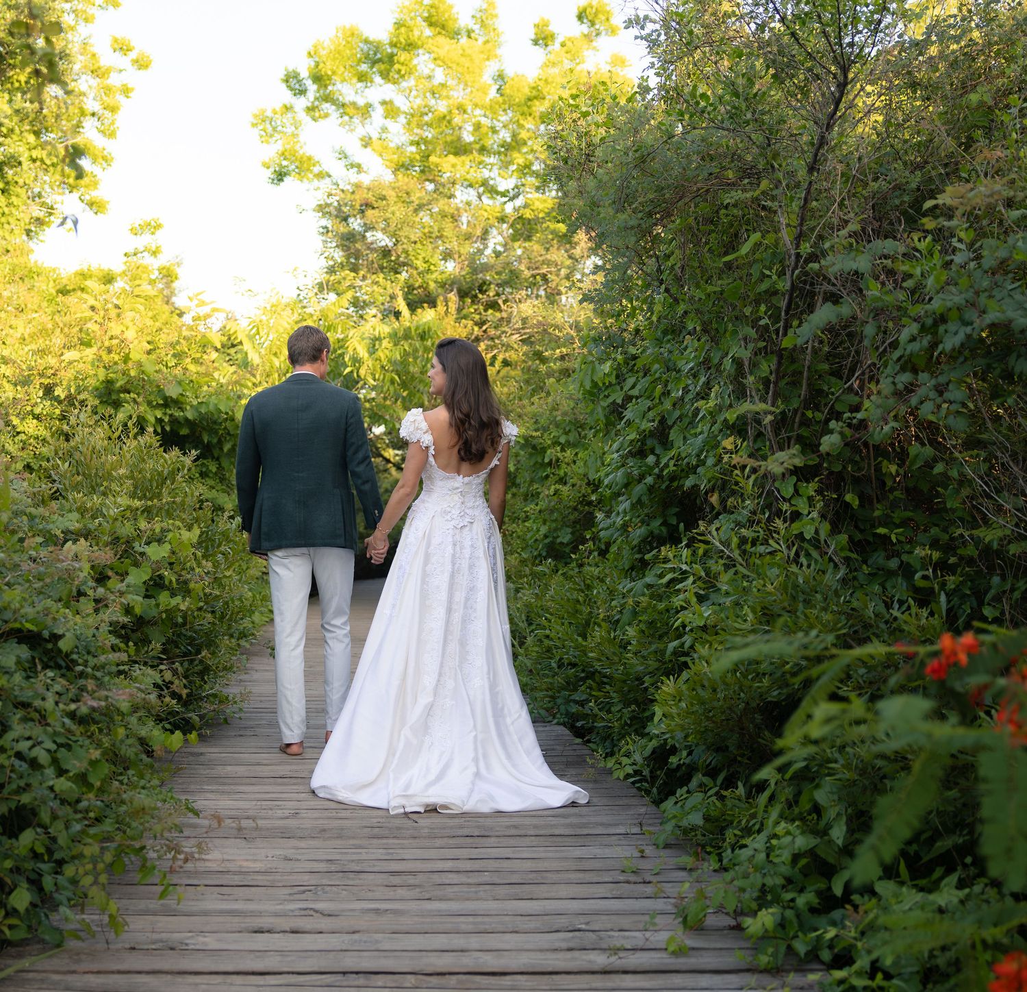 Bride and groom walking hand in hand along a wooden boardwalk surrounded by lush greenery.