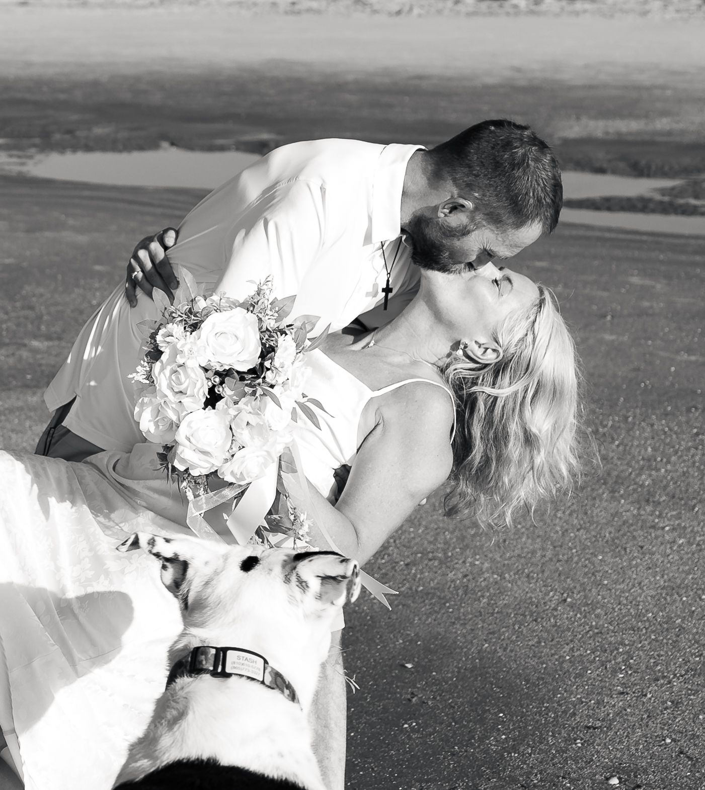 Black and white wedding photo of bride and groom sharing a dip kiss with a white dog at their feet outdoors.