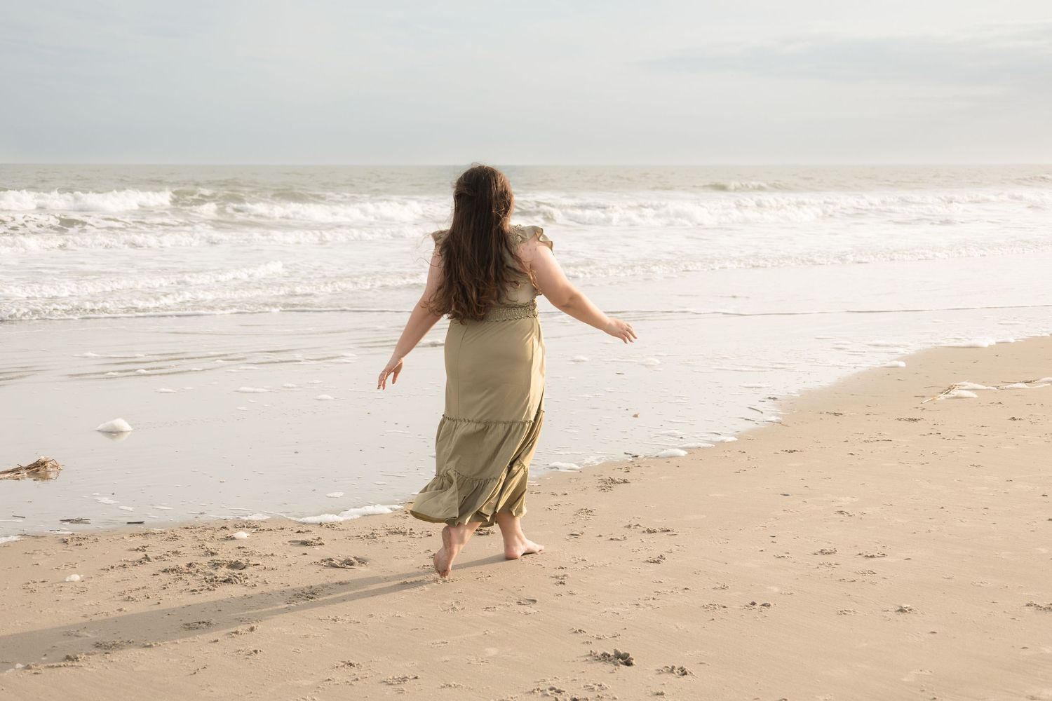 A person in a beige dress twirls joyfully on a sandy beach at sunset with waves in the background.